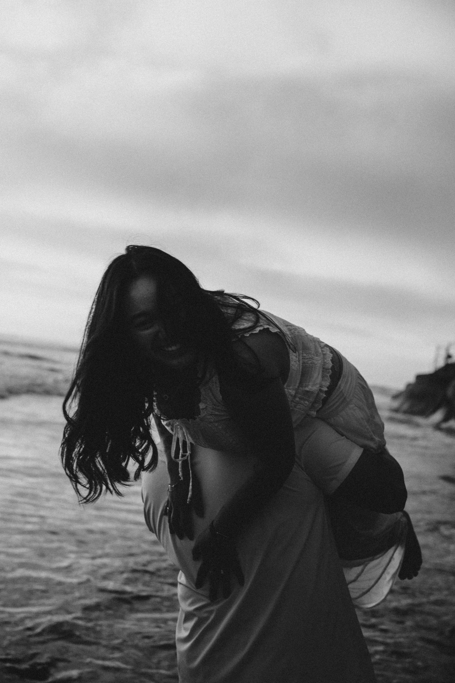 Black and white photo of laughing woman being carried on beach, hair blowing in wind with dramatic cloudy sky.