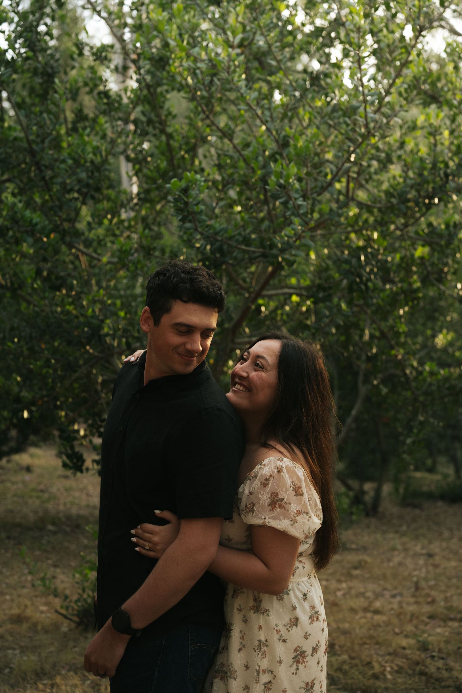 Smiling couple embracing warmly among lush green trees in a natural outdoor woodland setting.