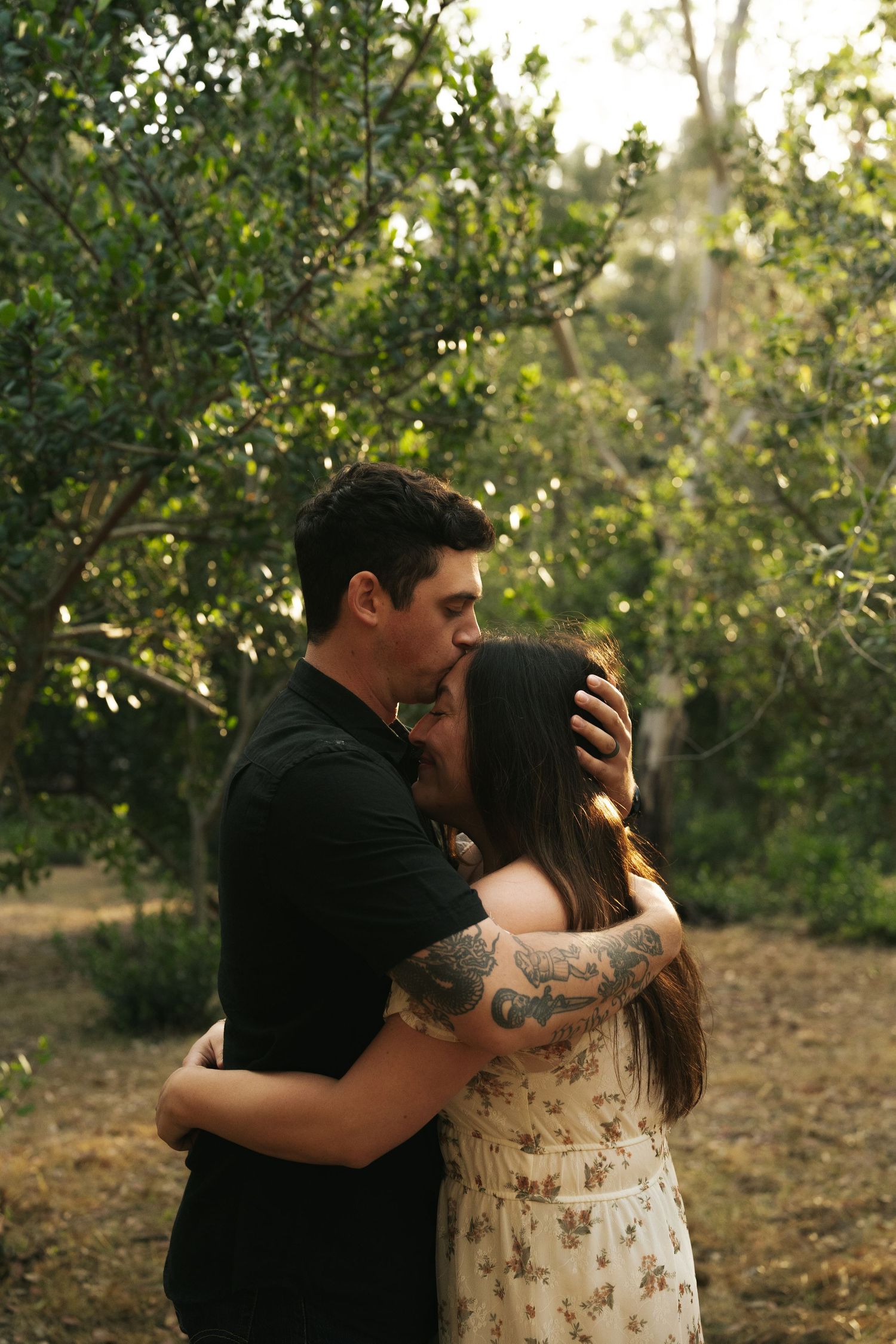 A tattooed couple shares a tender kiss outdoors, surrounded by lush green trees in warm natural light.