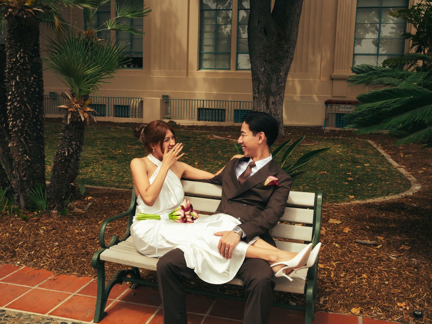 A bride and groom sit on a garden bench, sharing a romantic moment outside a classic building.
