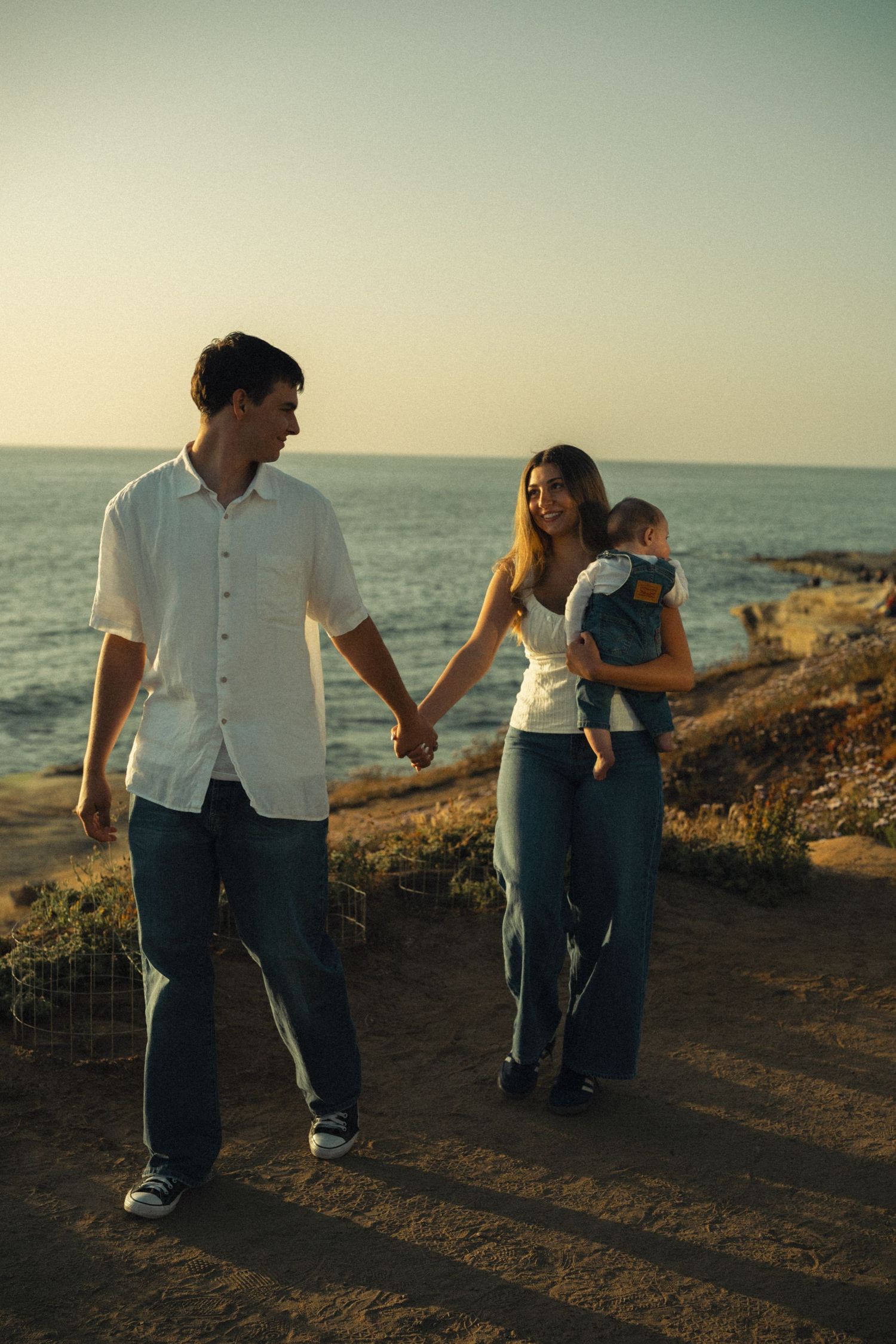 A young couple holds hands while walking along a coastal path at sunset, with the woman carrying a baby in a carrier.