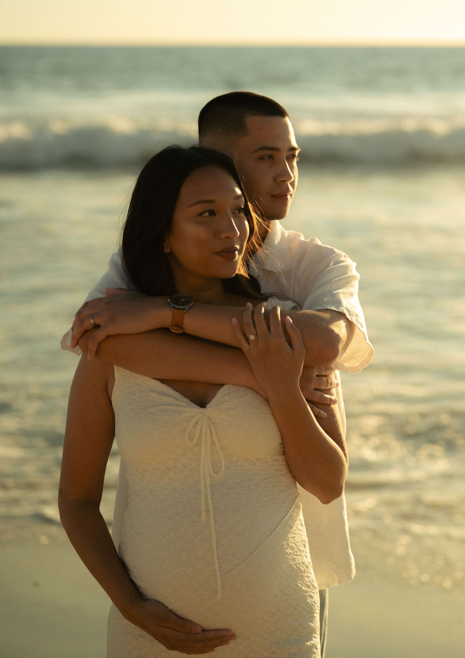 Man embraces pregnant woman from behind on beach at sunset maternity shoot.