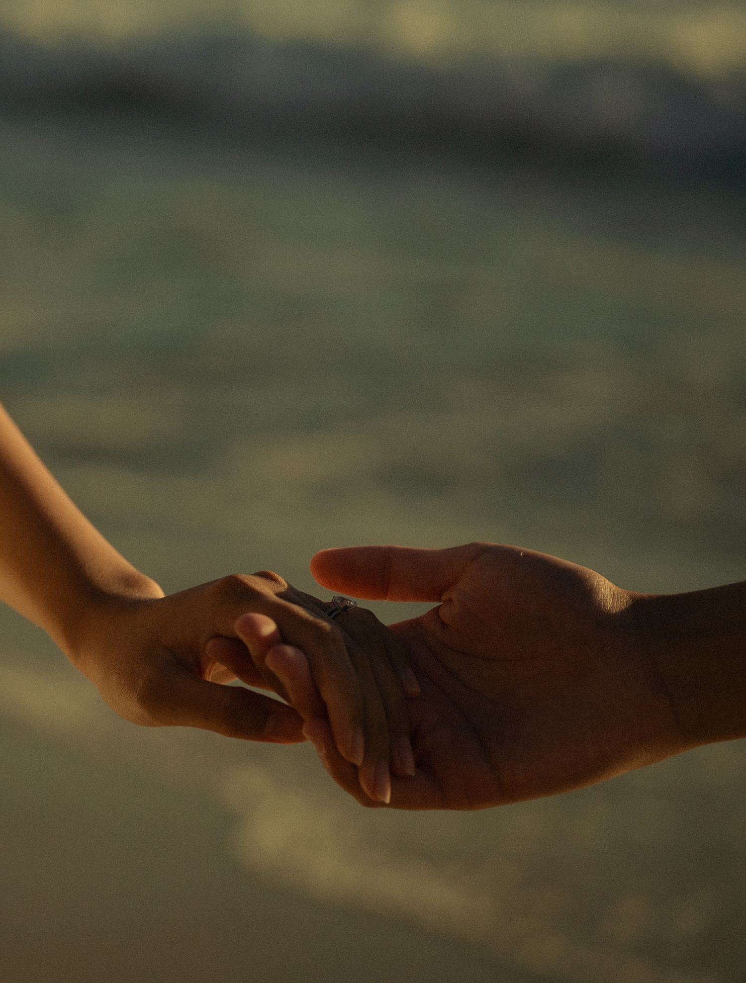 Two hands gently touching at a beach during golden hour, symbolizing romance and connection by the ocean.