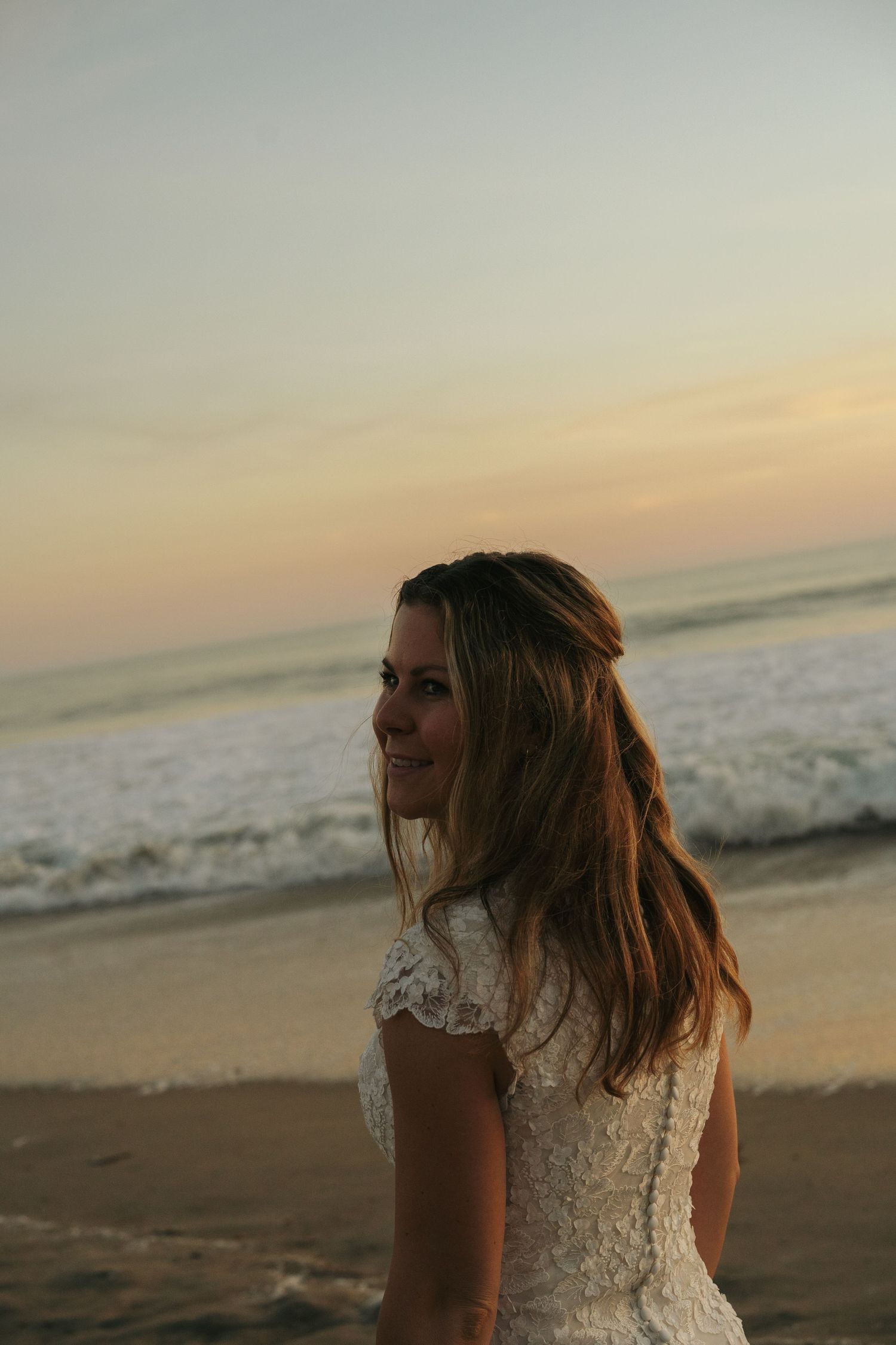 Smiling bride in white lace dress on beach at sunset with ocean waves.
