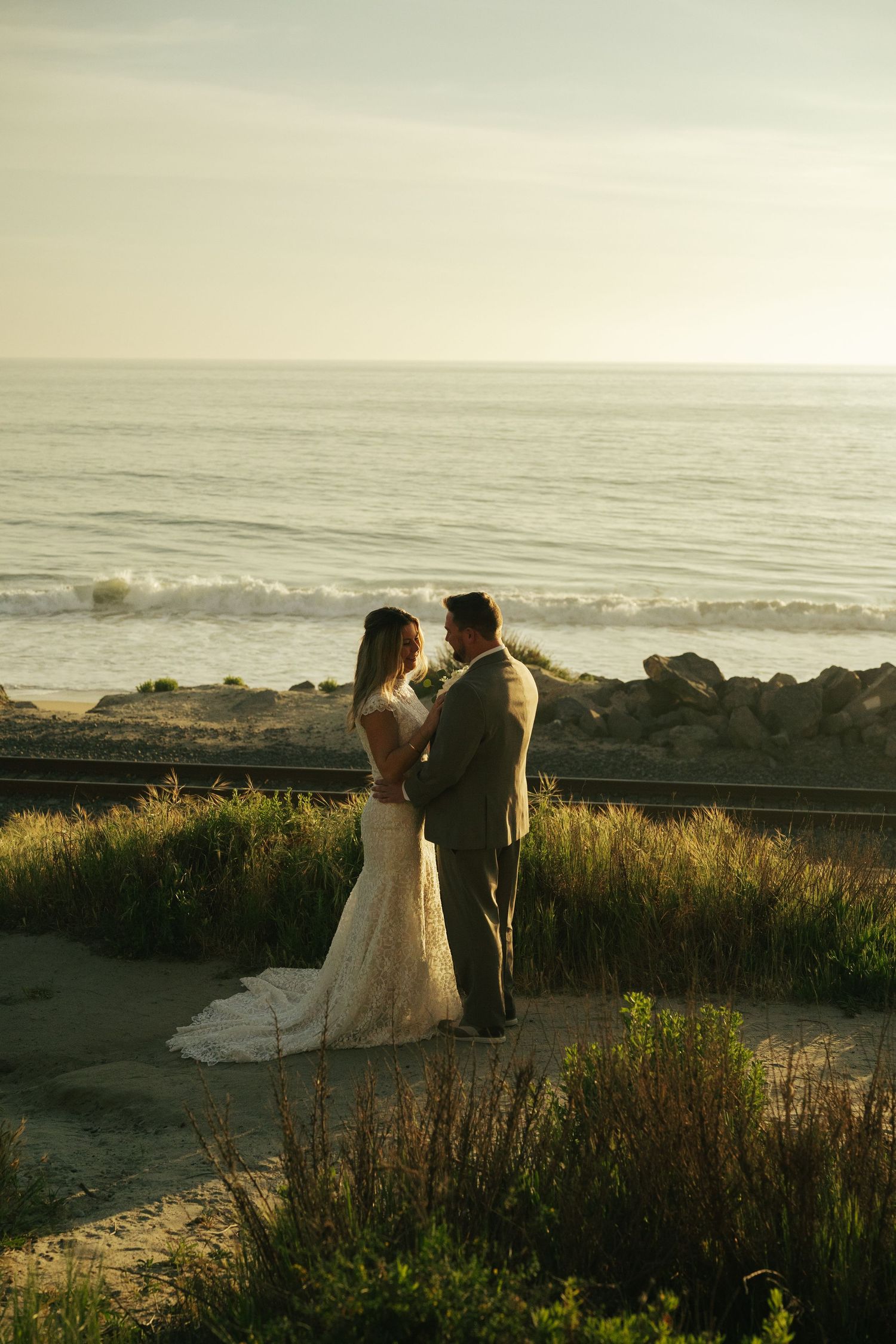 Bride and groom stand facing each other near ocean railroad tracks at golden hour sunset beach wedding.