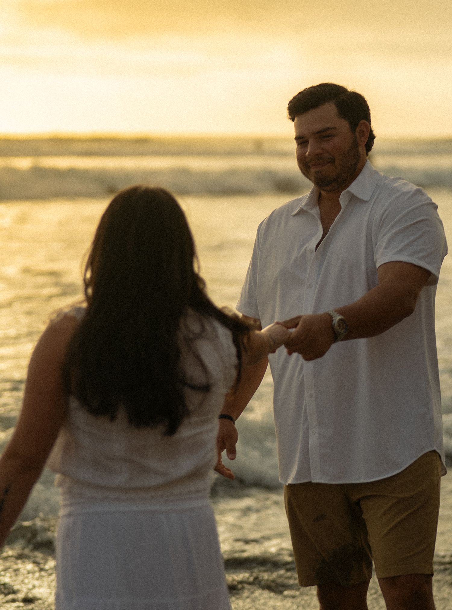 Couple holding hands on a beach at golden sunset, man in white shirt and khaki shorts, woman in white dress.