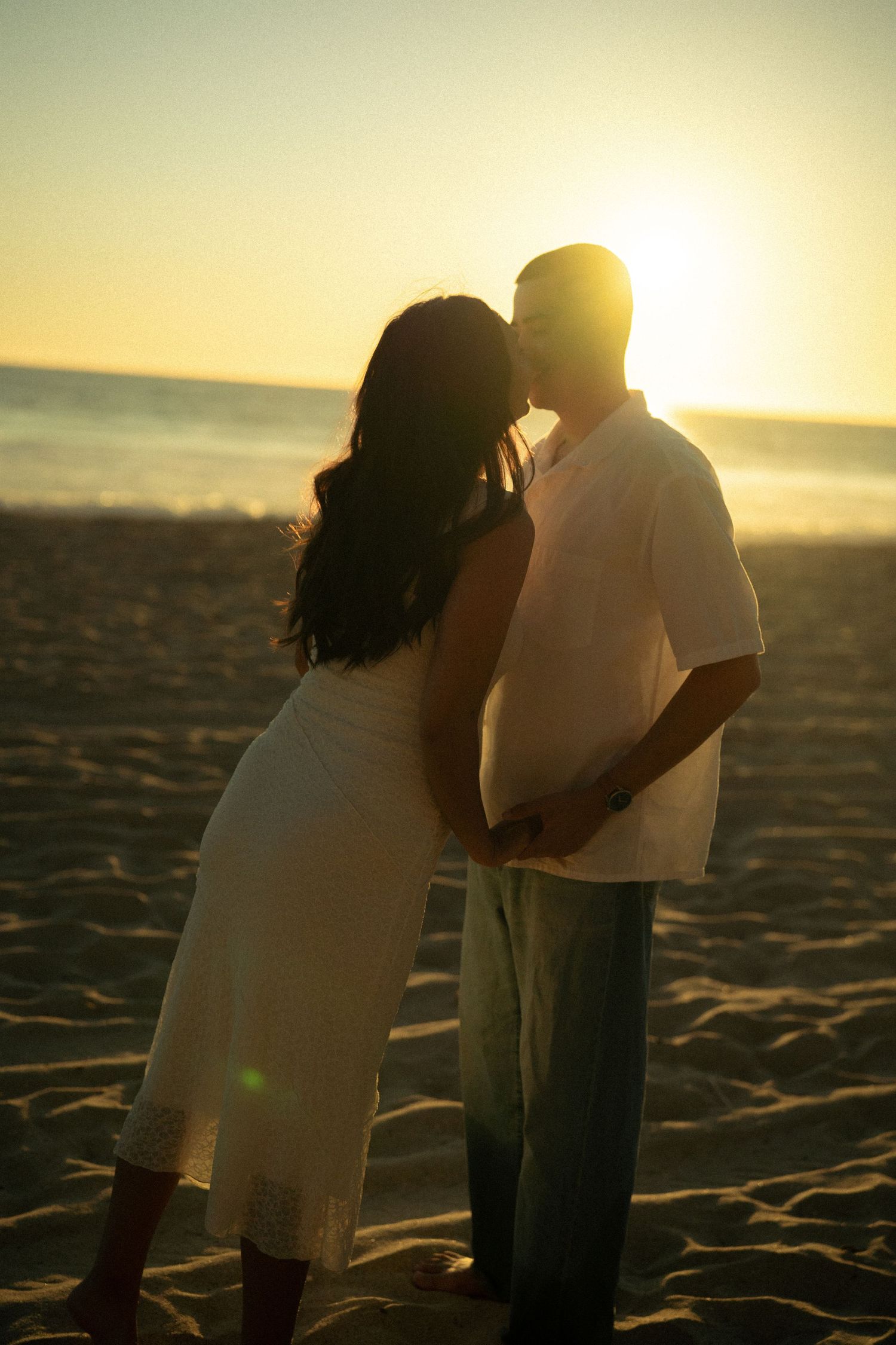Couple sharing a kiss at sunset on beach, silhouetted against warm golden sky.