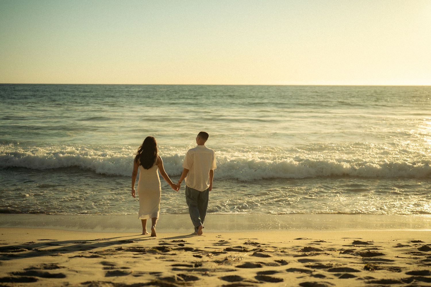 Couple holding hands walking along a sandy beach toward ocean waves at sunset.