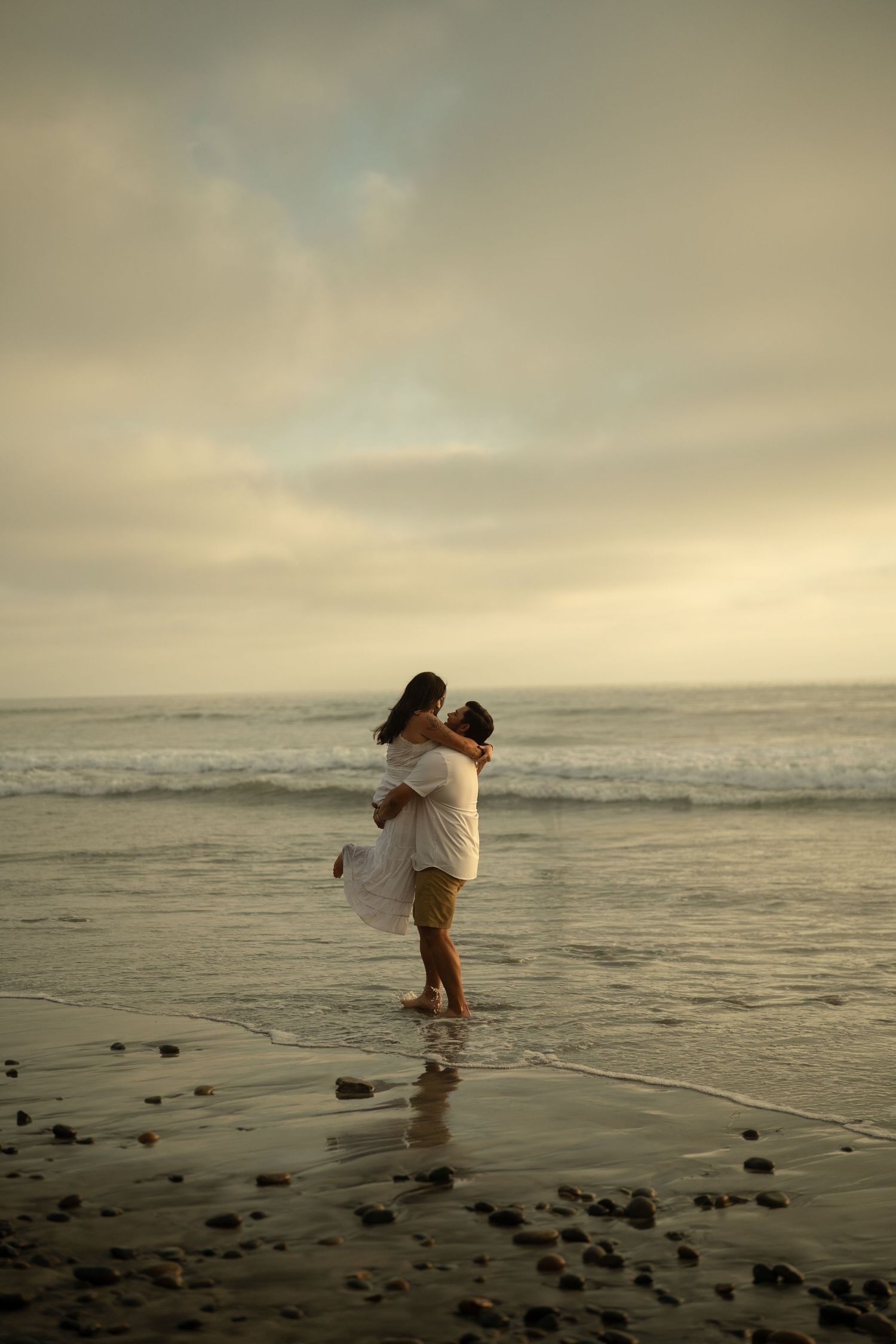 A couple shares a romantic embrace on a misty beach at sunset, man lifting woman in white dress by the ocean waves.