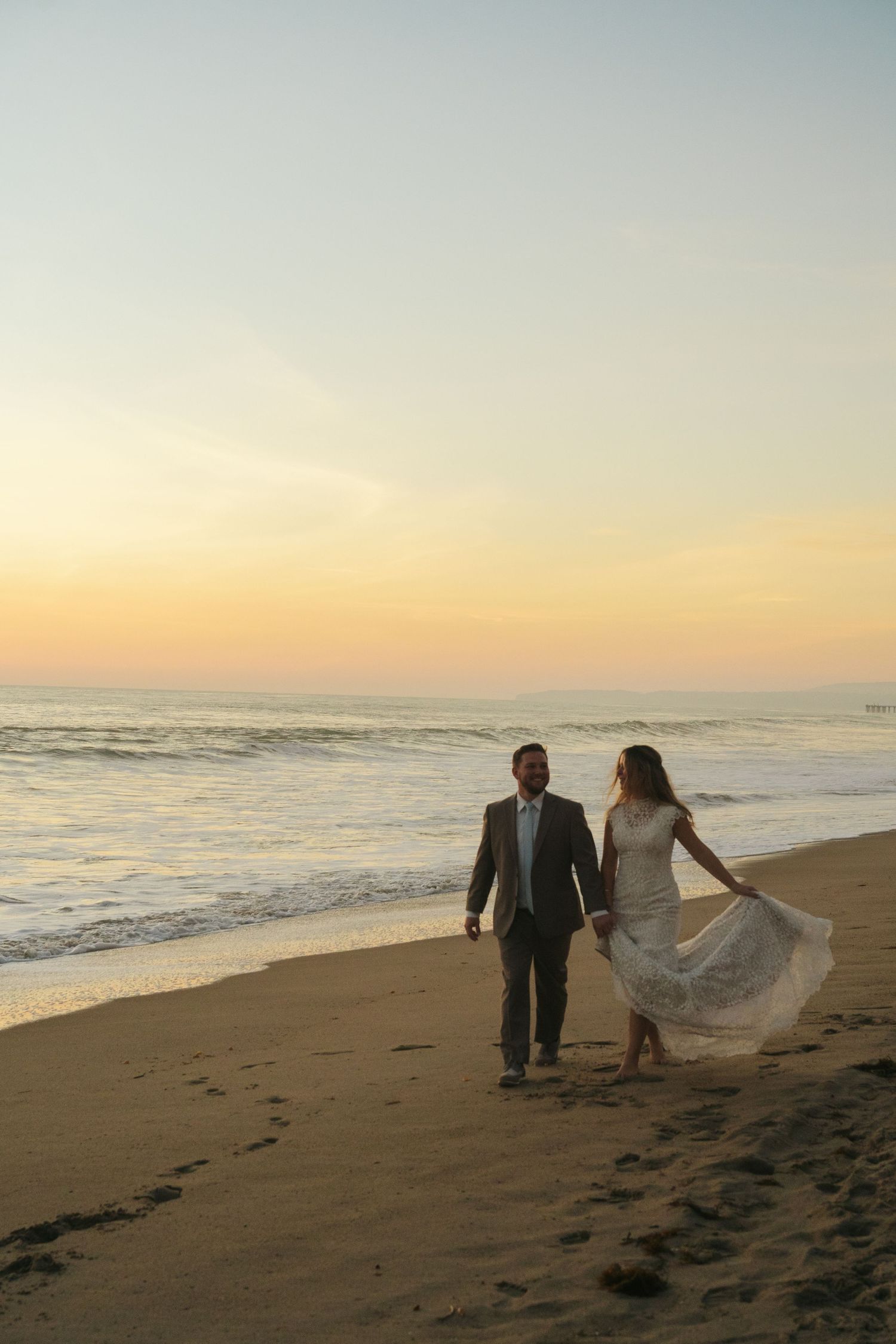 Bride and groom walk hand-in-hand along a sandy beach at sunset, waves gently rolling in behind them.