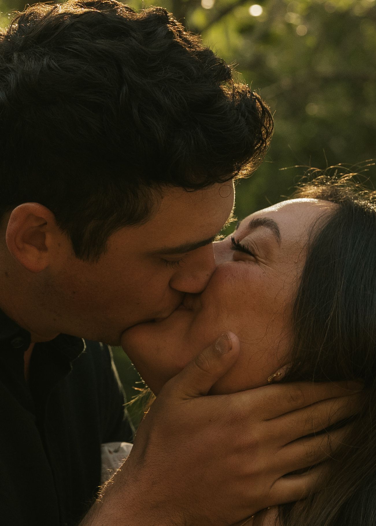 A couple sharing a tender kiss outdoors in warm golden light, with the man gently cradling the woman's face.