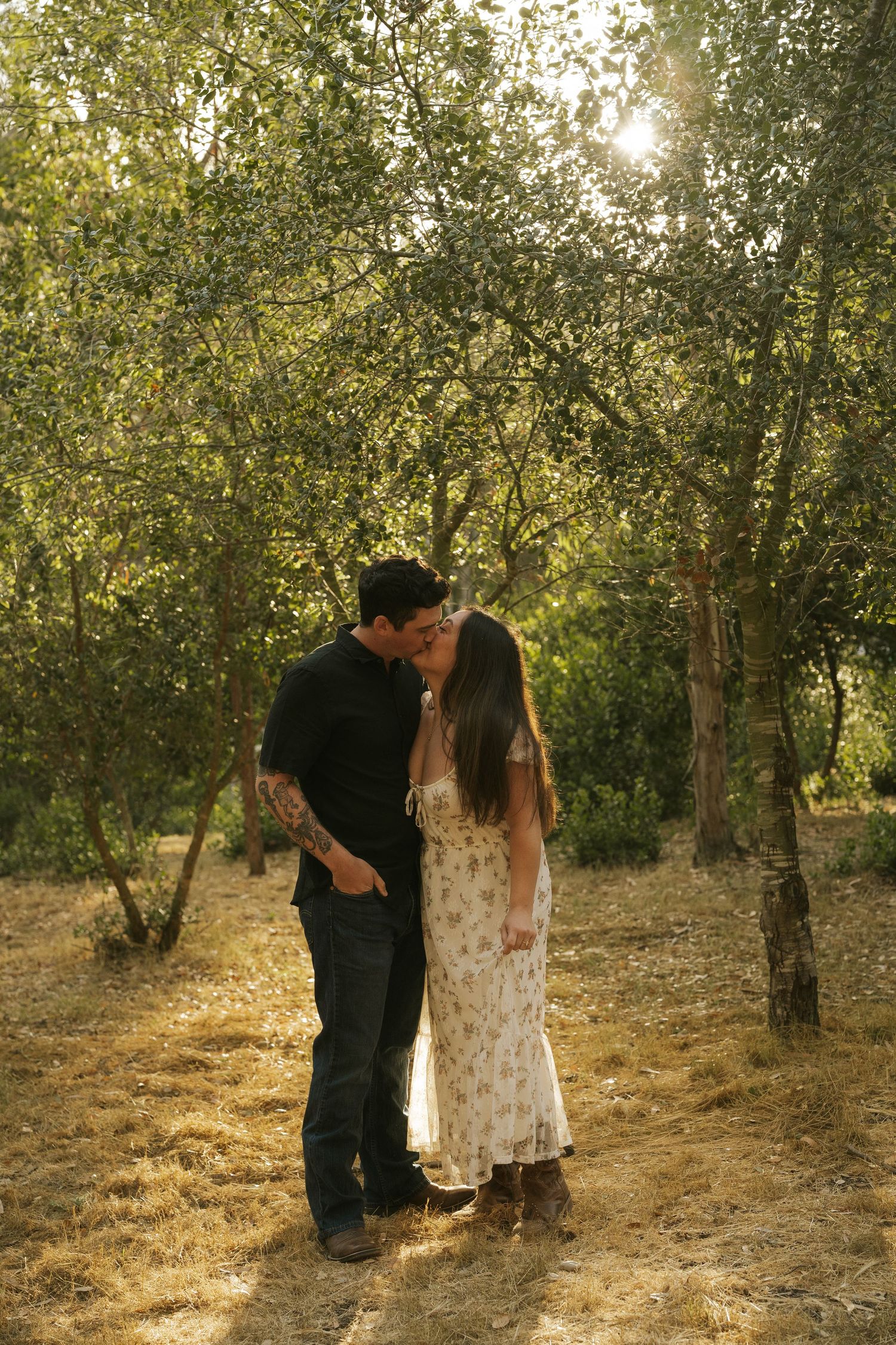 Couple kissing tenderly among sun-dappled trees in a peaceful outdoor setting.