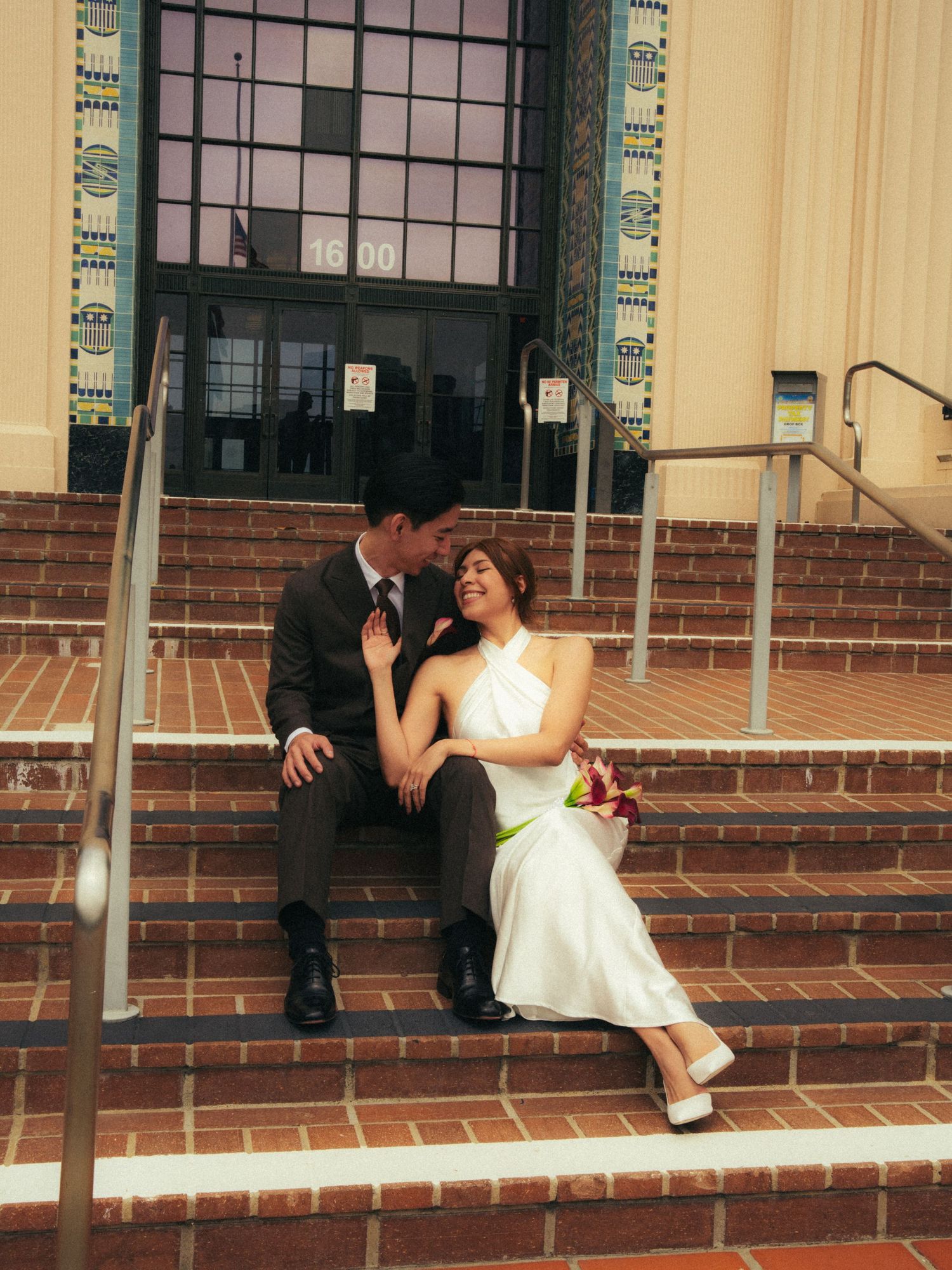 A bride and groom sit romantically on brick steps outside a building, sharing an intimate moment in wedding attire.
