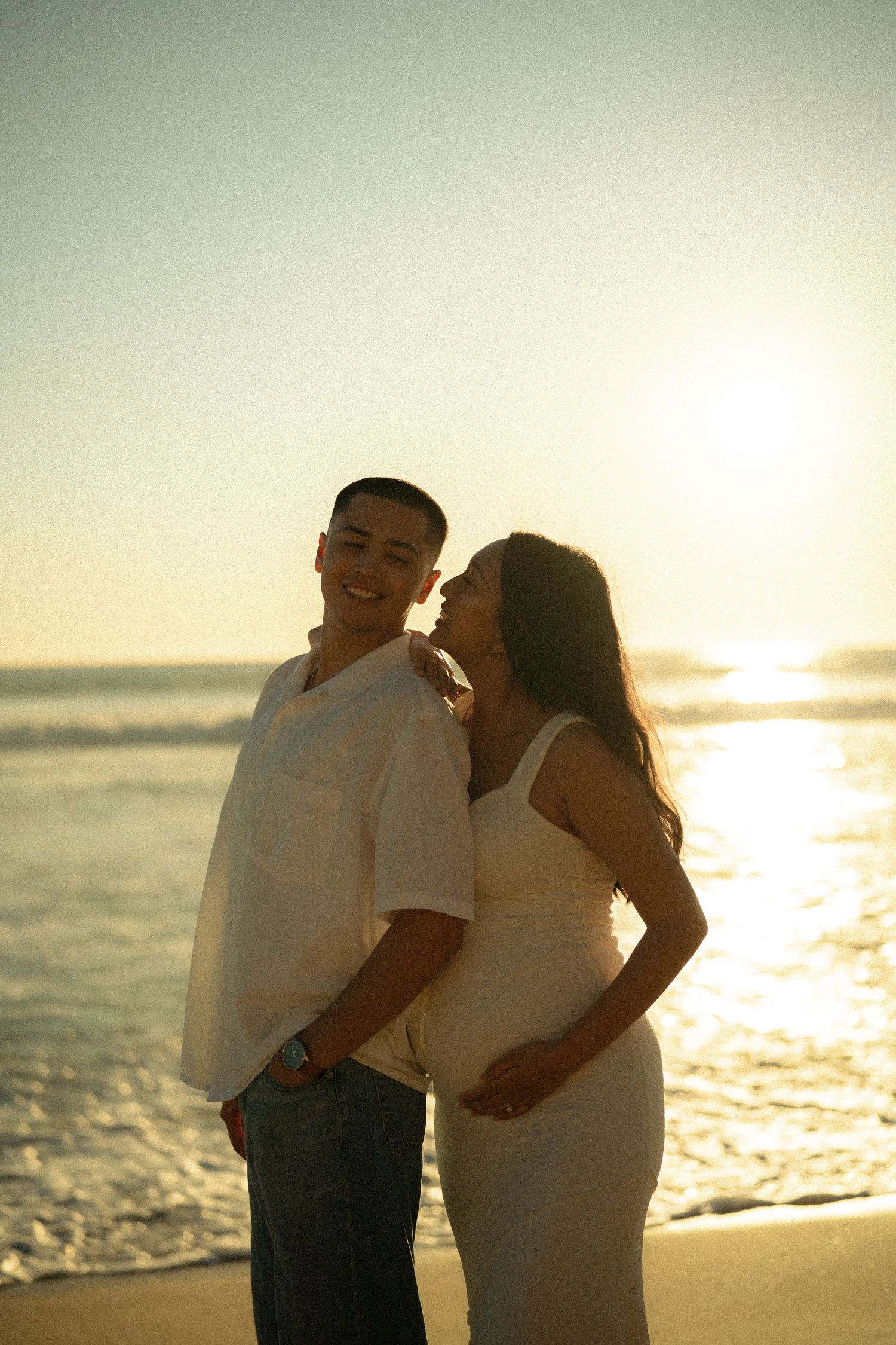 Couple embracing on beach at golden sunset, pregnant woman in white dress leaning on smiling partner by ocean waves.