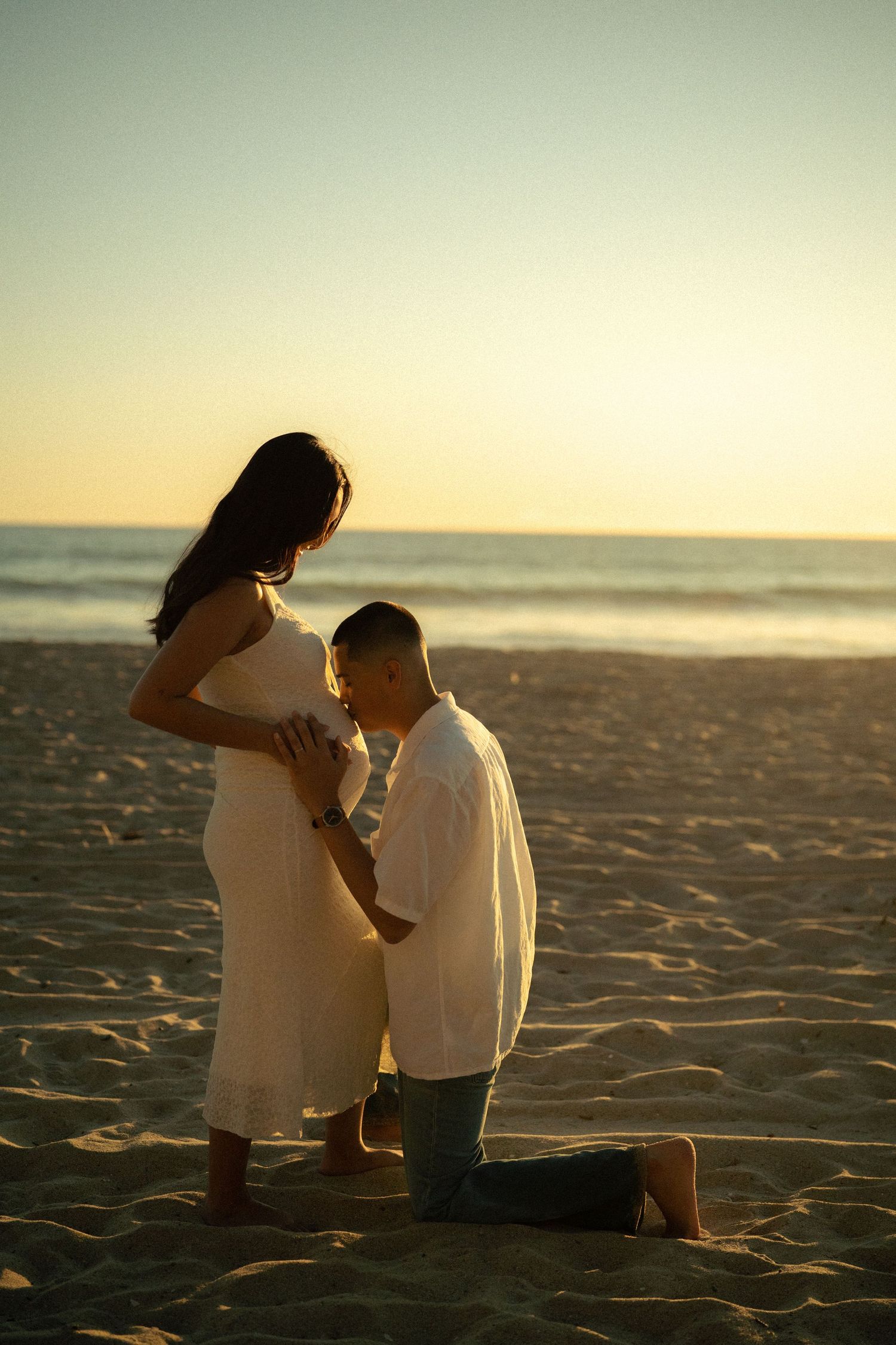 Man kneeling on sandy beach kissing pregnant partners belly at sunset by ocean shoreline.