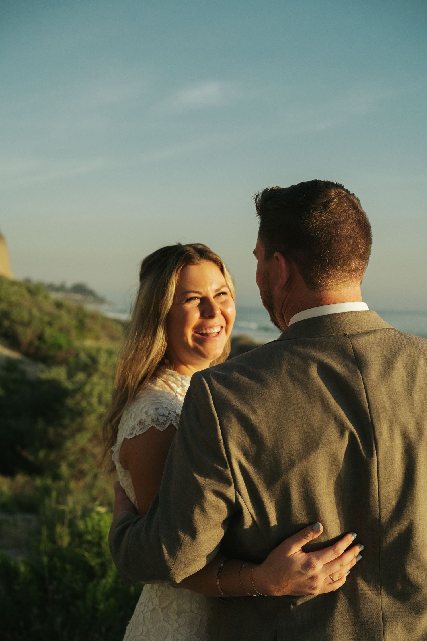 Smiling bride in lace wedding dress embracing groom outdoors with ocean and cliffs in golden sunset background.