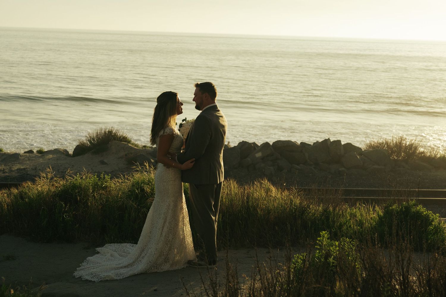 A couple in wedding attire shares a romantic moment by the ocean at sunset, surrounded by coastal greenery.