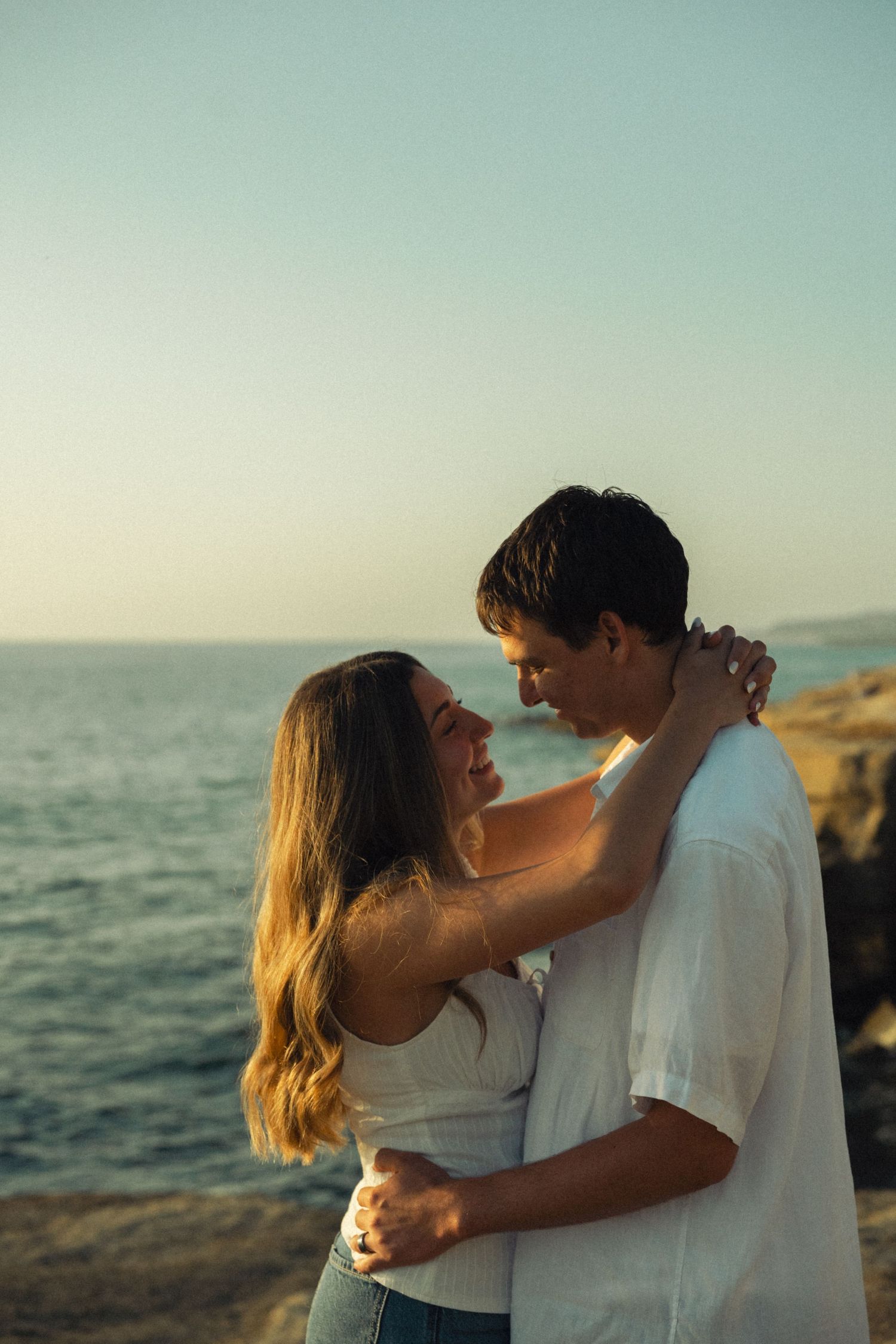 Romantic couple embracing and smiling at each other on a rocky ocean cliff at sunset.