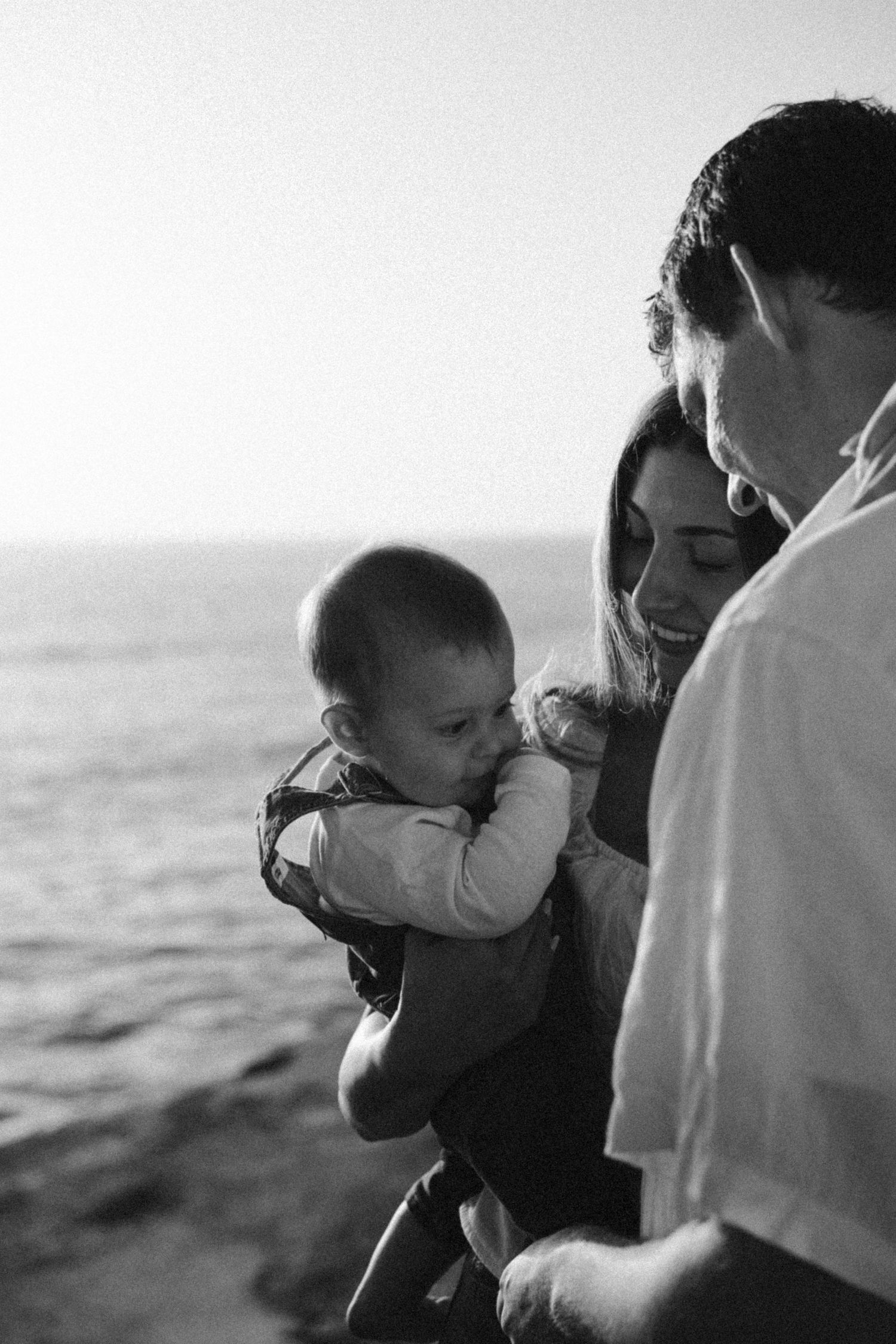 Black and white photo of loving parents cradling their infant baby by the sea.