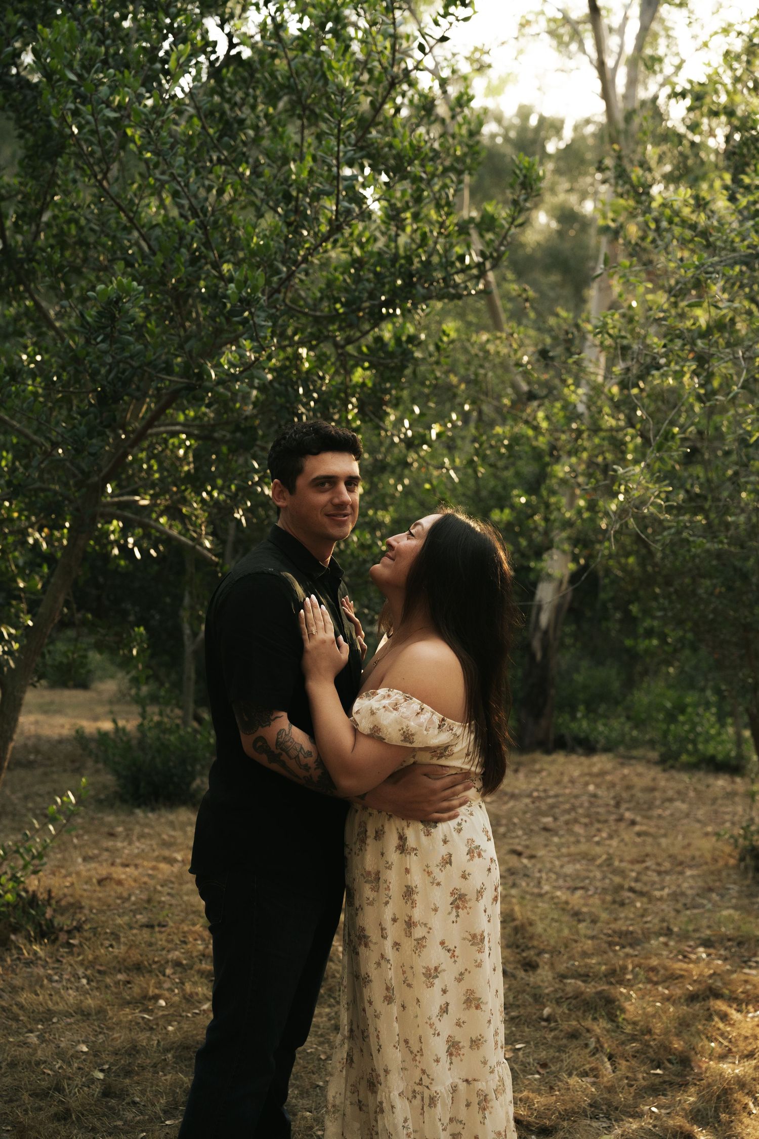 Couple embracing in a lush green forest, woman in floral dress laughing joyfully while man in black shirt smiles.