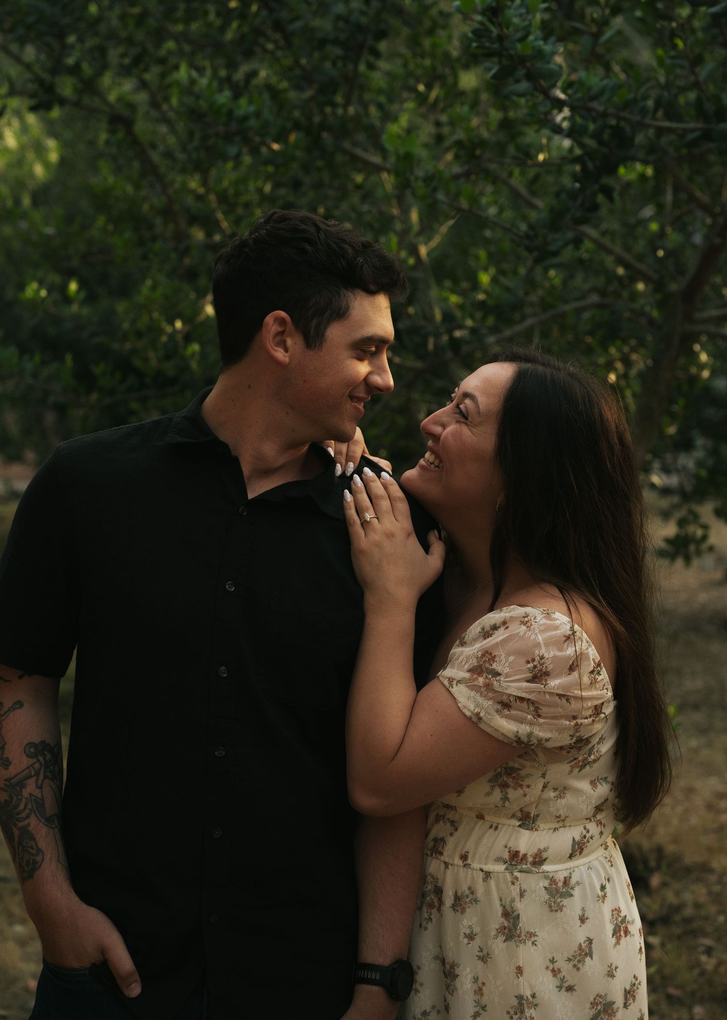 Couple smiling at each other intimately in a dark wooded outdoor setting at dusk.
