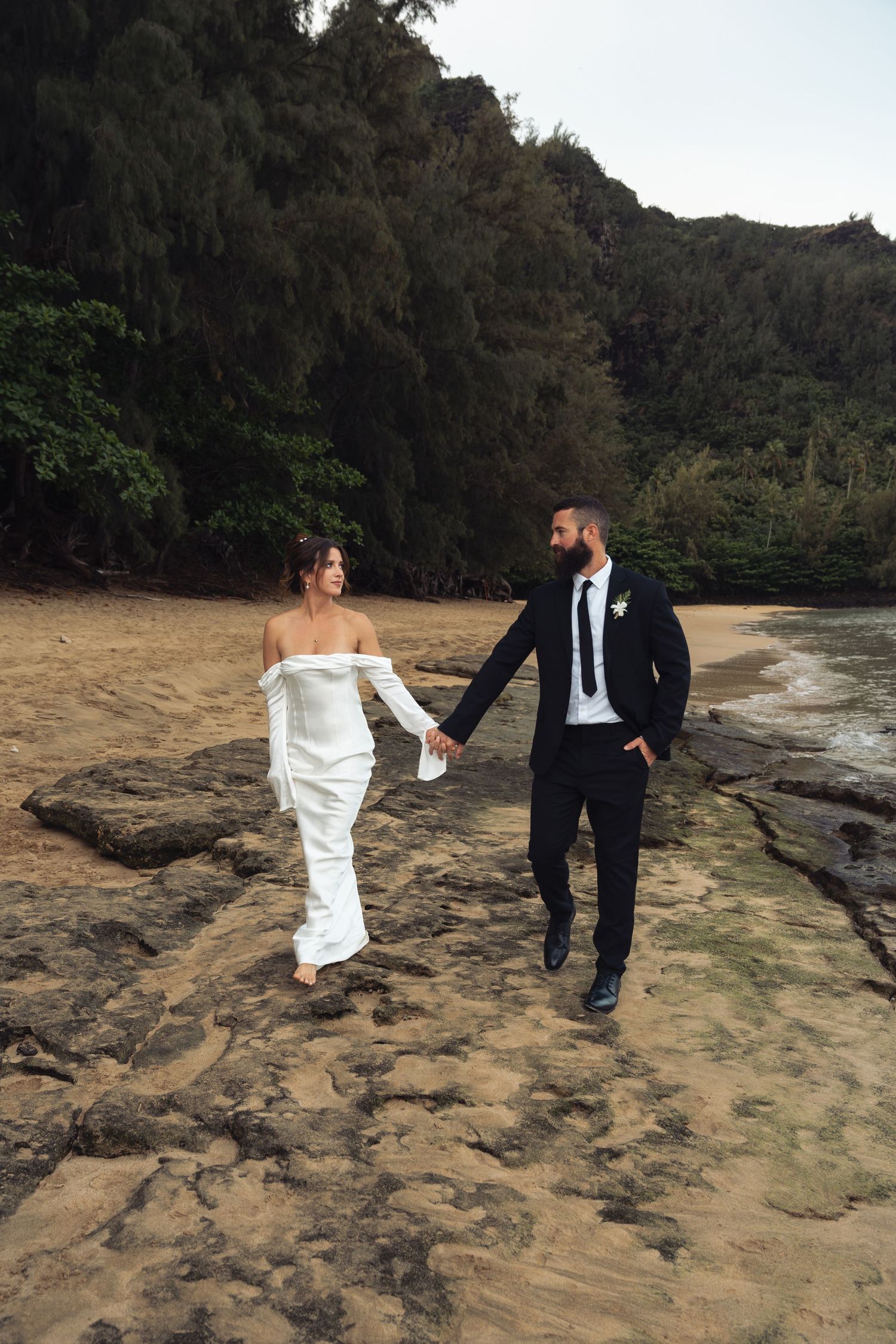 Newlyweds walk hand in hand along rocky beach coastline in formal wedding attire.