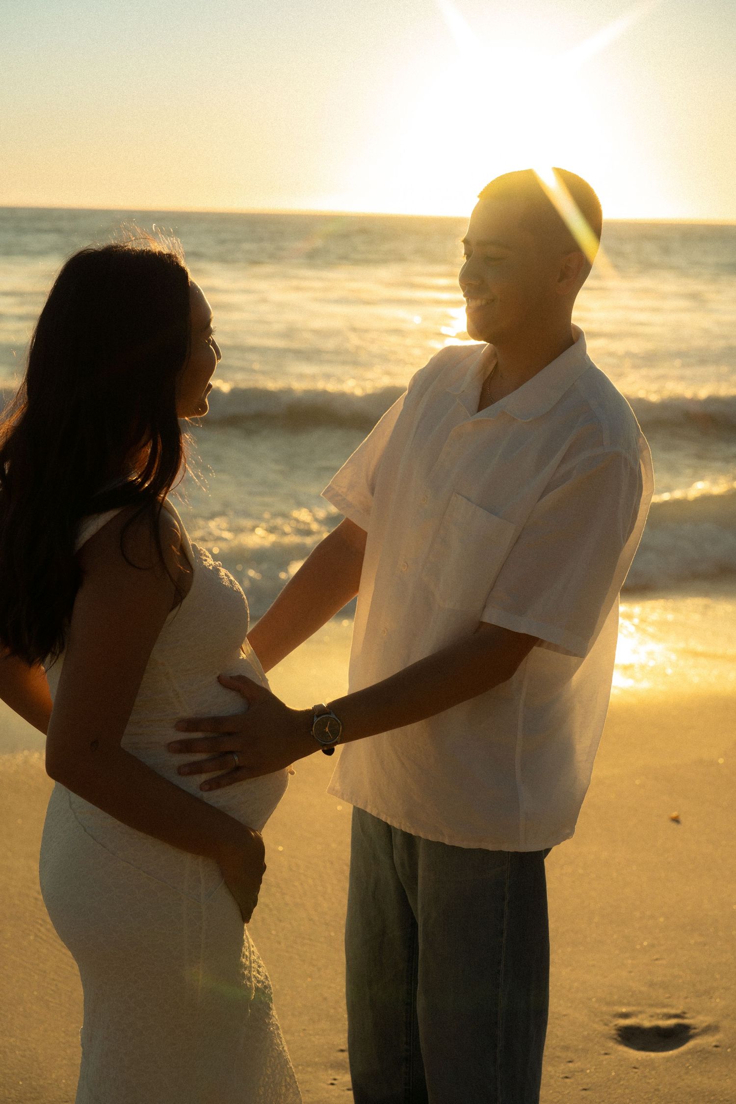 Couple holding pregnant belly together on beach at golden sunset with ocean waves behind them.