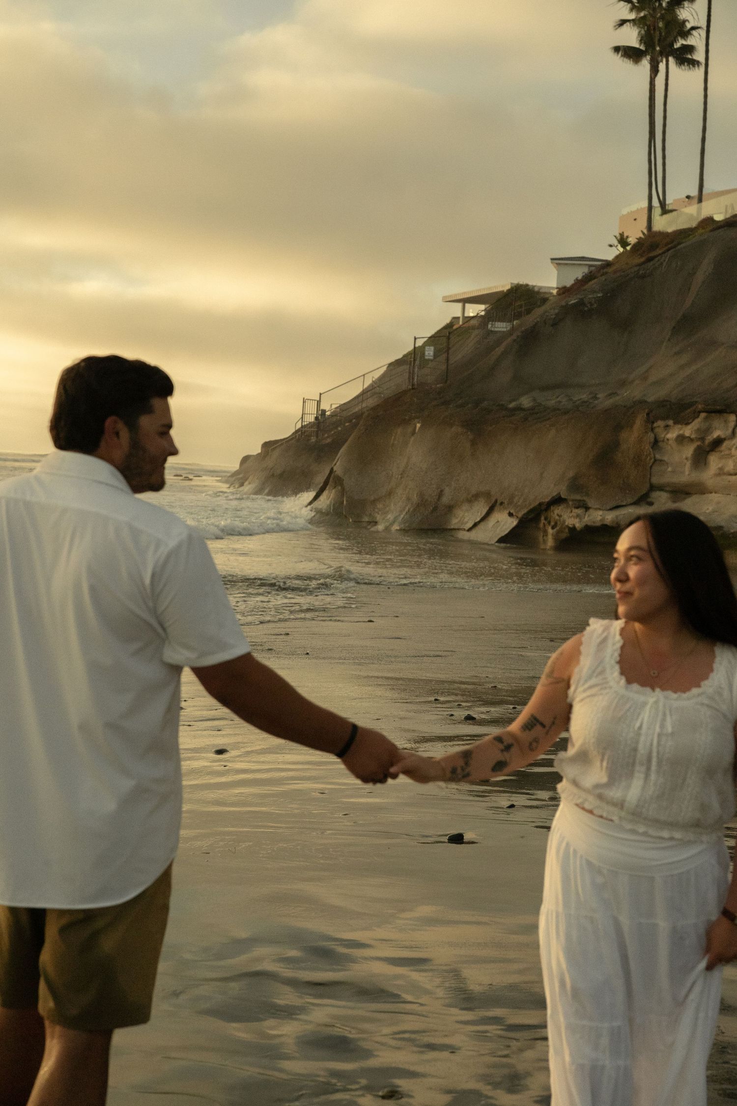 Couple holding hands on a sandy beach at sunset with ocean waves and rocky cliffs in the background.