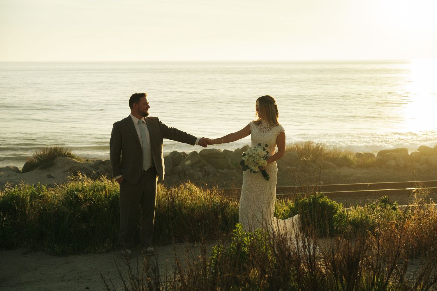 Bride and groom hold hands facing each other with golden ocean sunset backdrop in romantic wedding portrait.