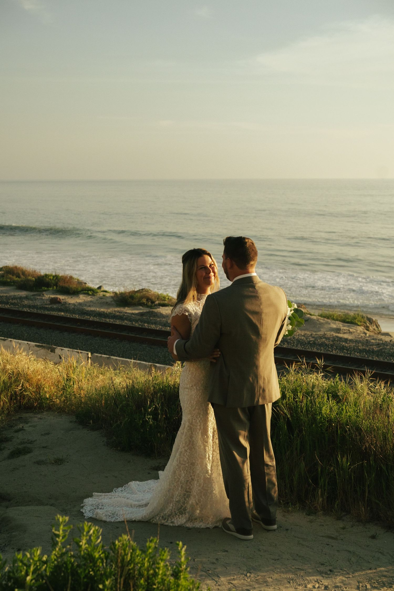 Bride and groom share a romantic moment overlooking the ocean at sunset in a coastal wedding photo.