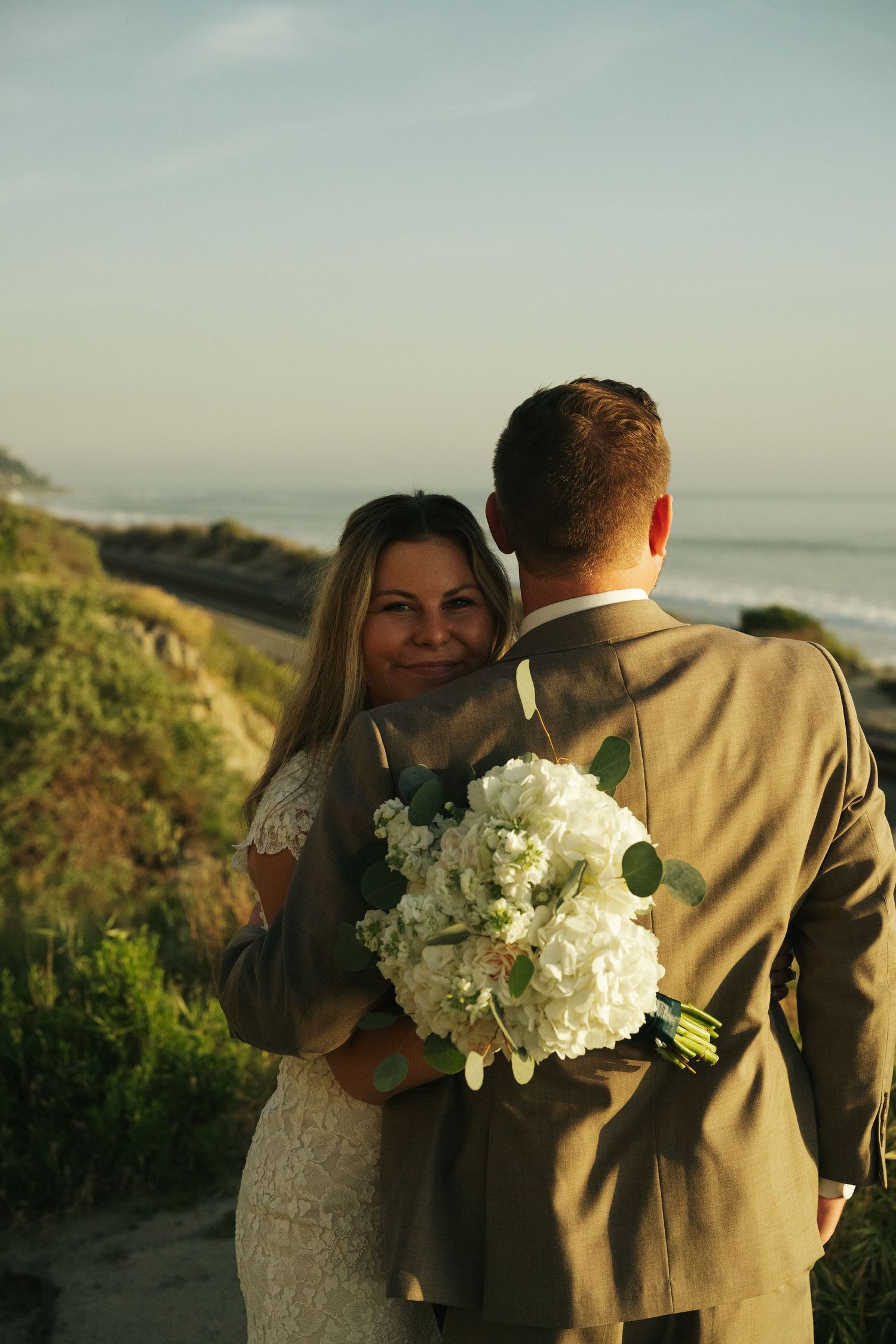 Bride holding white hydrangea bouquet embraces groom in tan suit with ocean view at golden hour.