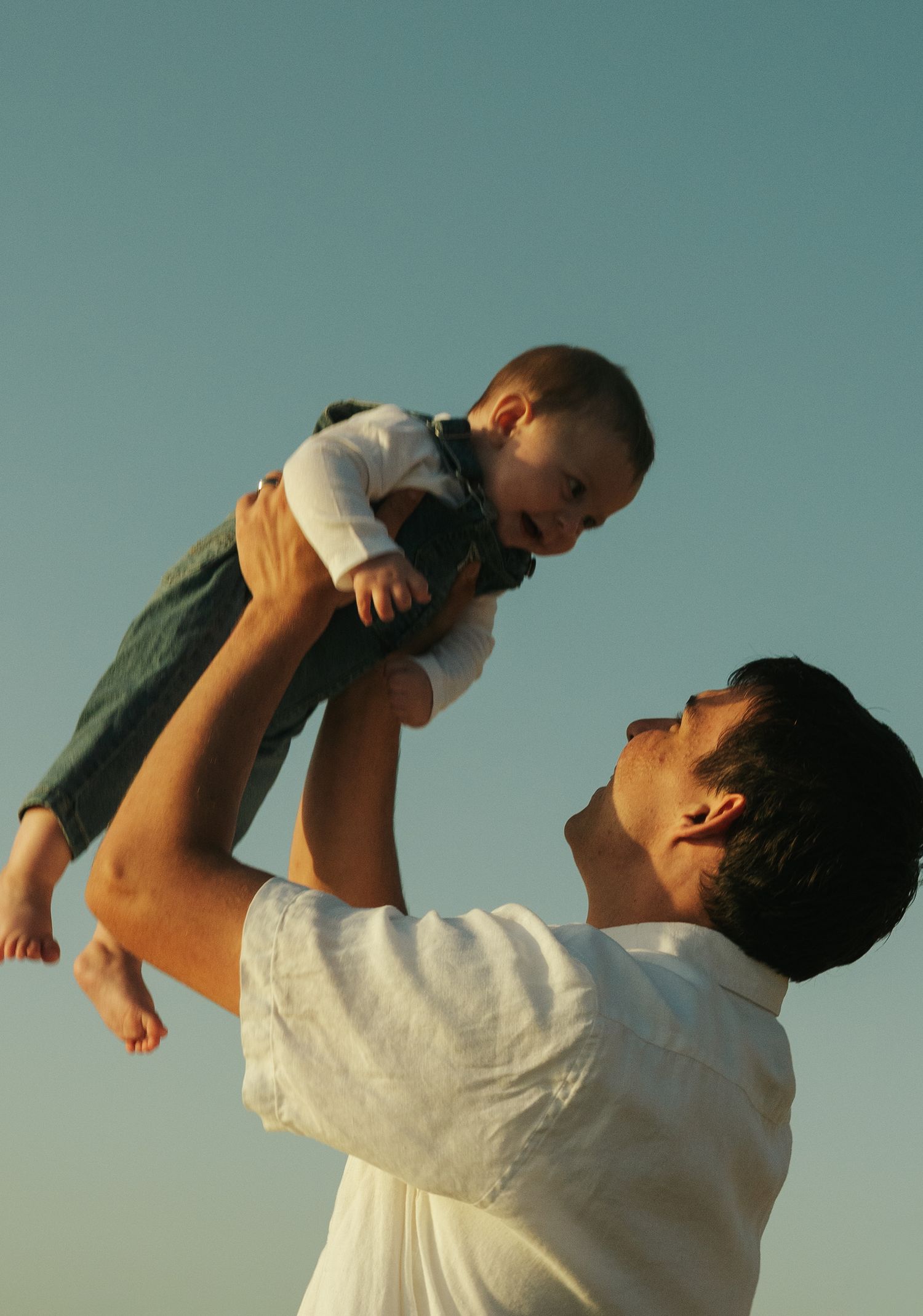 Joyful father lifting laughing baby high in the air against clear blue sky outdoors.