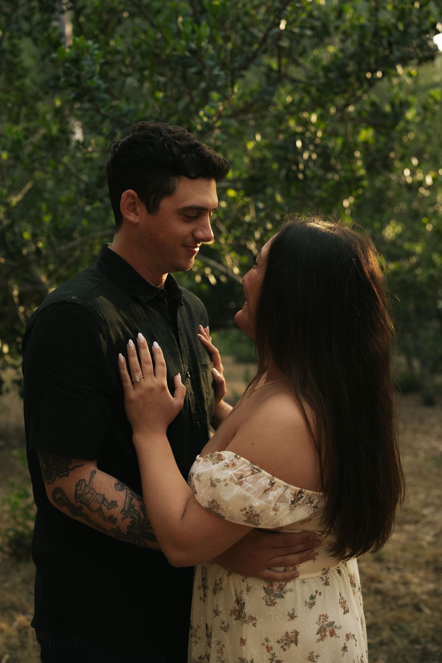 A couple gazes lovingly at each other in a garden, woman in floral off-shoulder dress touching mans chest.