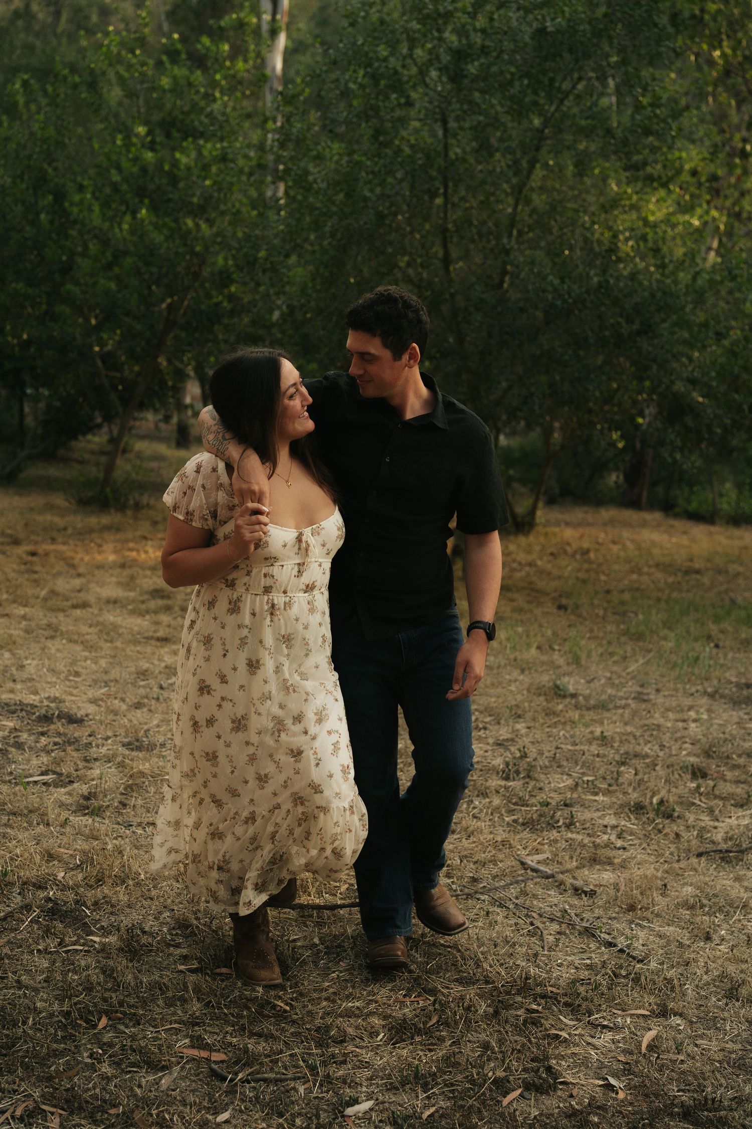 A couple walks arm in arm through a green orchard at dusk, sharing a warm intimate glance.