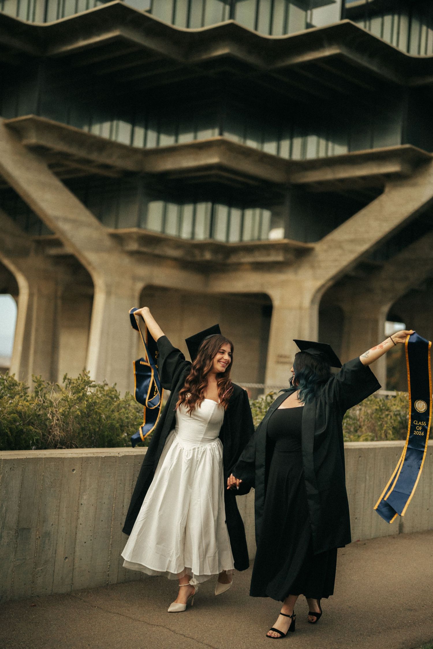 Two graduates in black caps and gowns celebrate outdoors, raising their stoles joyfully in front of a modern building.