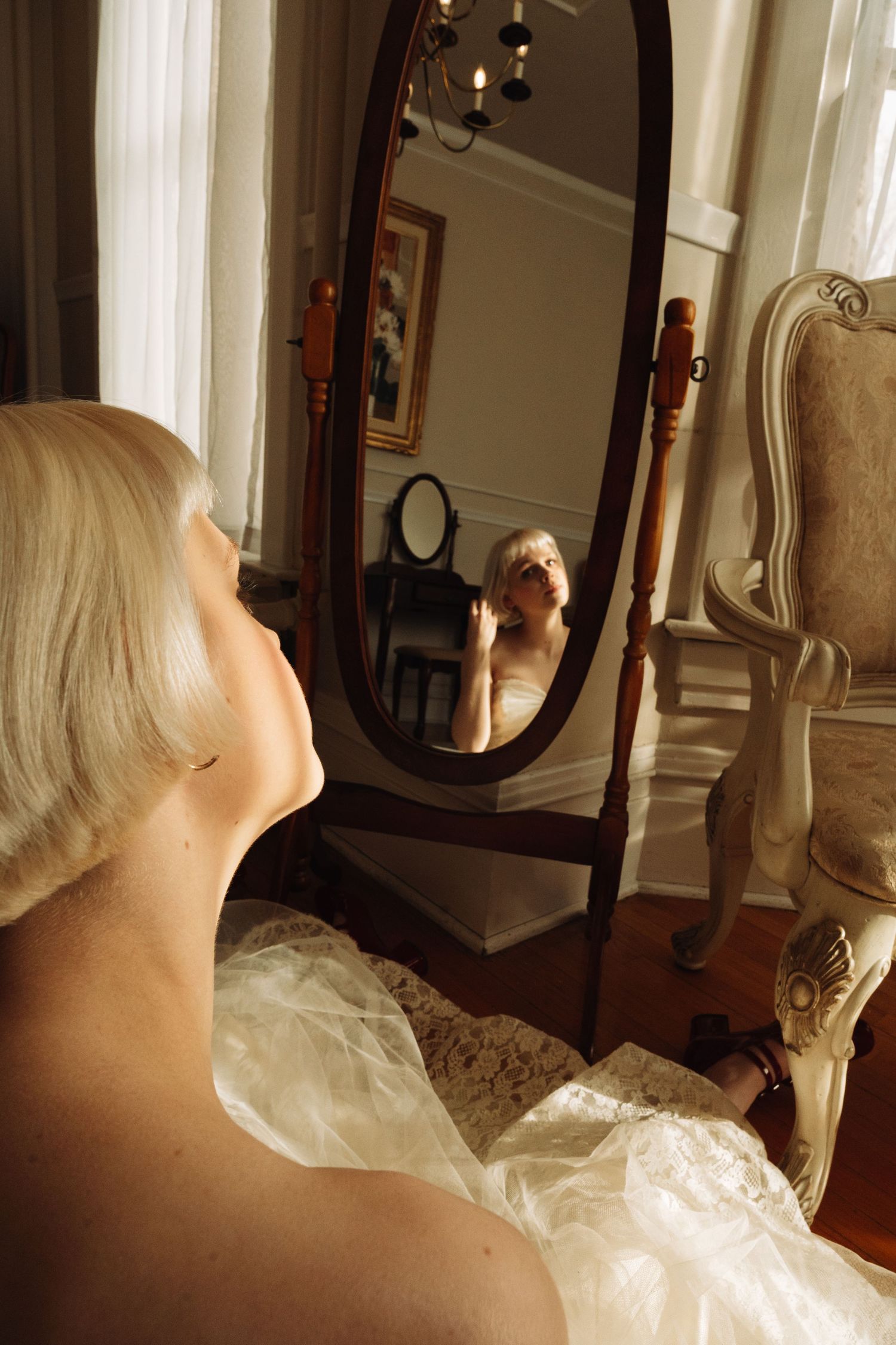 A reflection in an antique oval mirror shows someone in a white dress in an elegant room.
