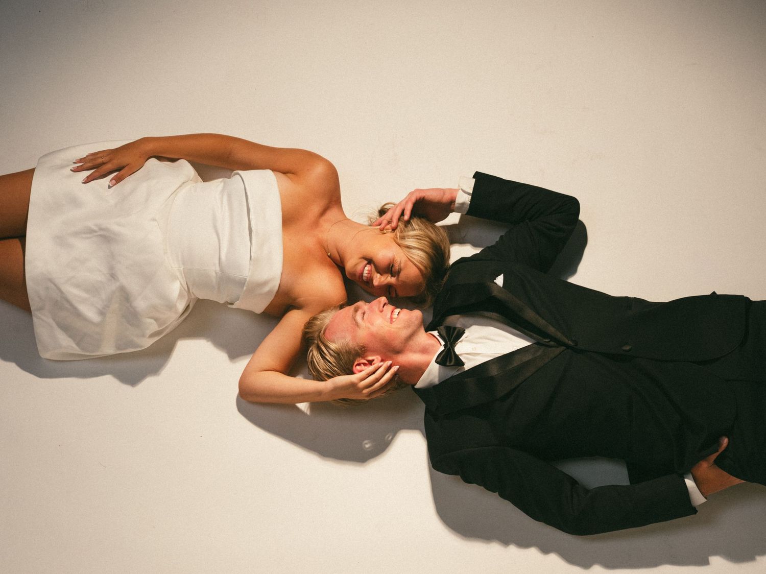 Bride in white strapless gown and groom in black suit lying on floor face to face in romantic wedding photo.