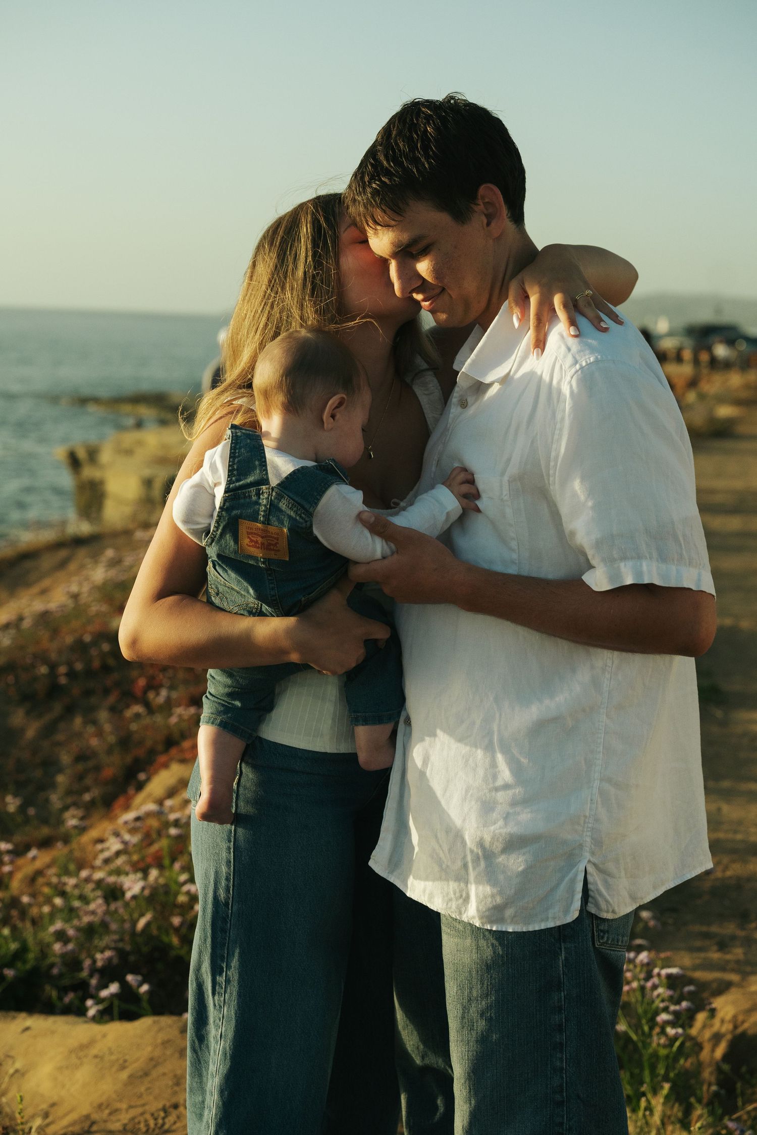 A young couple sharing a kiss while holding their baby near a rocky ocean coastline at sunset.