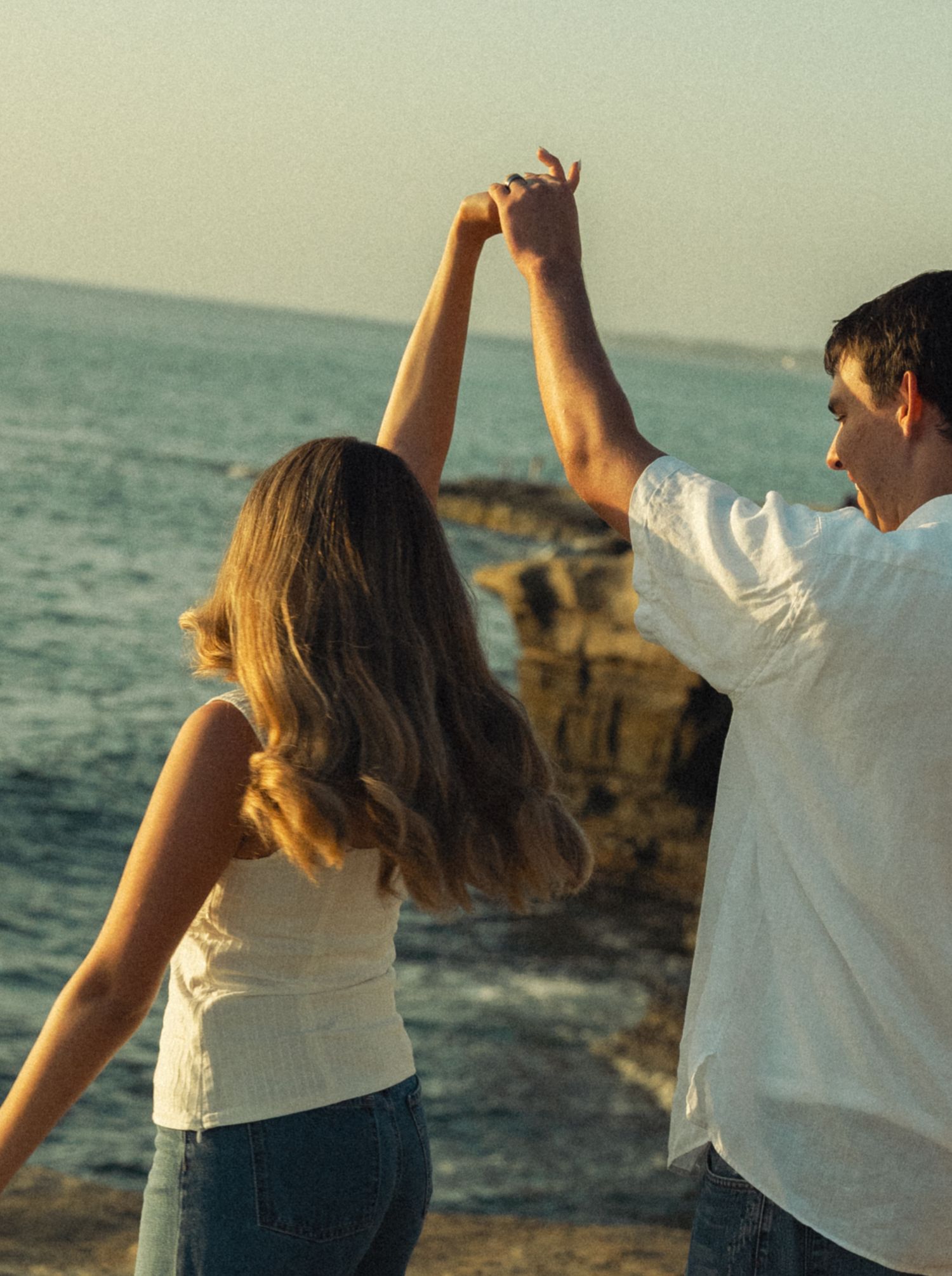 Couple dancing together holding hands raised by rocky ocean shore at sunset.