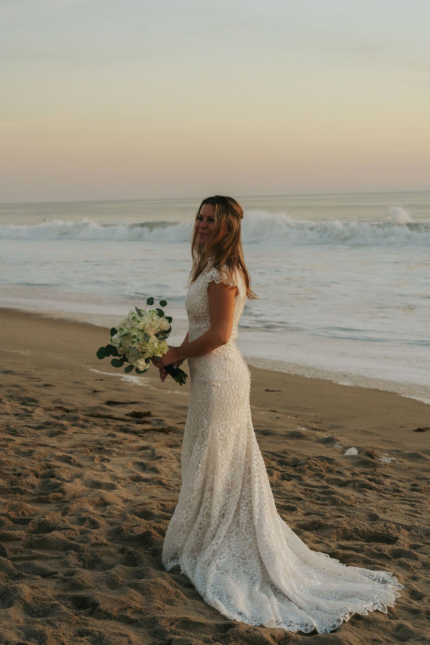 Bride in elegant lace wedding gown with train standing on sandy beach at sunset holding white bouquet.