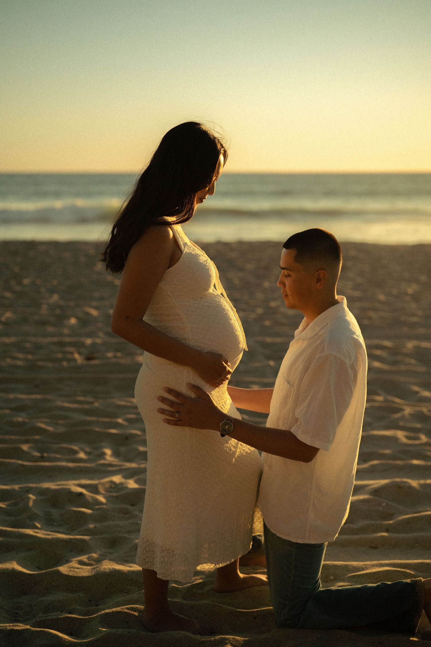 Man kneels on beach at sunset touching pregnant woman's belly in maternity photo.