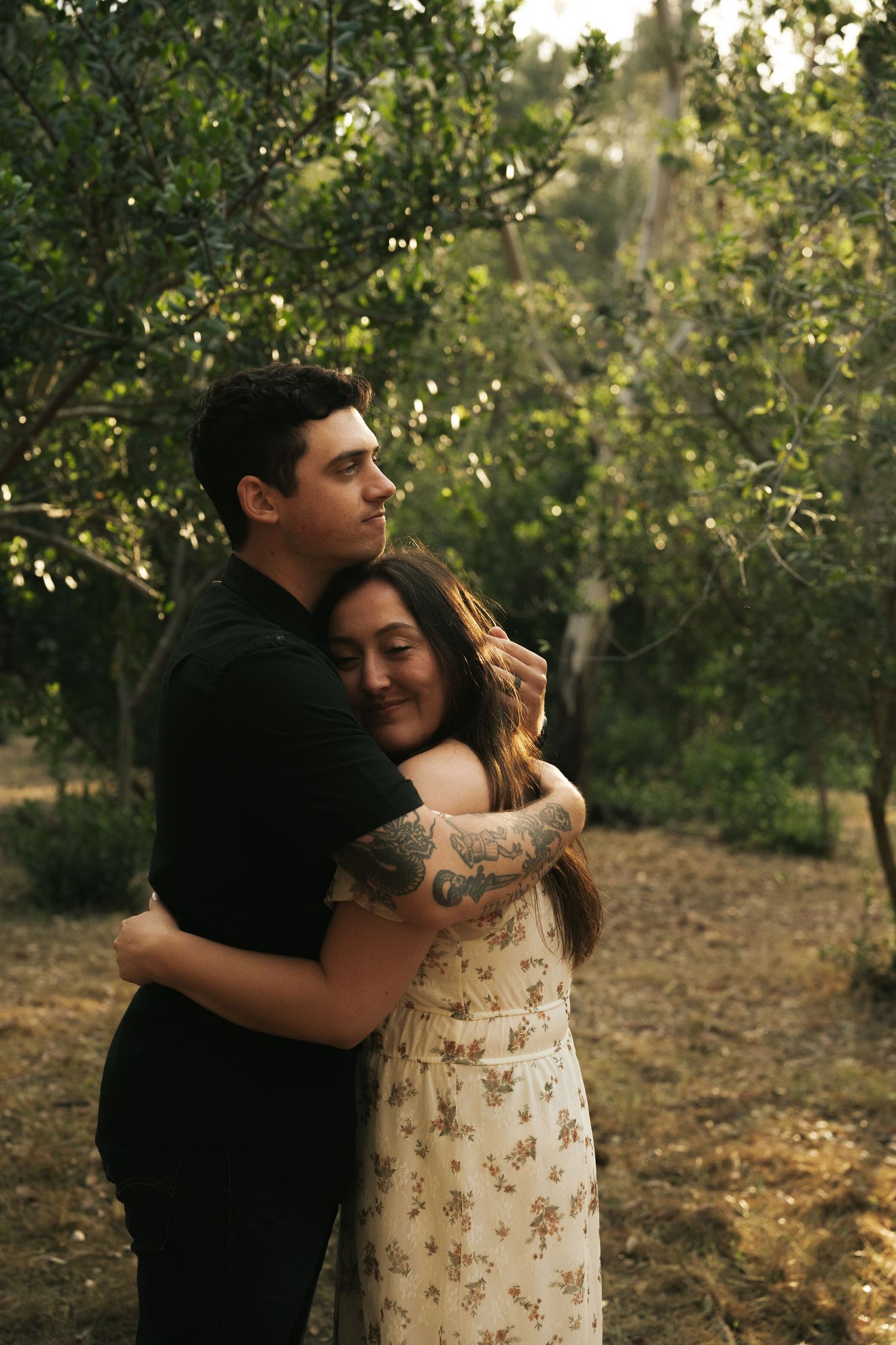 A couple embraces warmly among lush green trees, the woman smiling as the tattooed man holds her close.