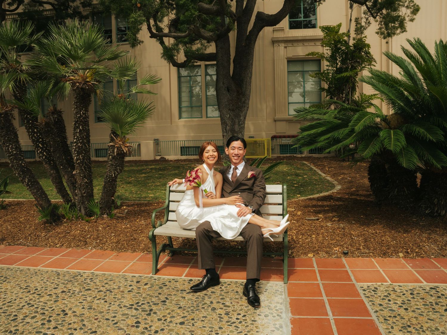 A happy Asian couple in wedding attire sits on a bench outdoors, surrounded by palm trees and a building.