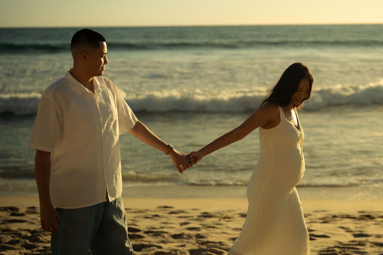 Pregnant couple holding hands on a sandy beach at sunset, wearing white outfits with ocean waves in the background.