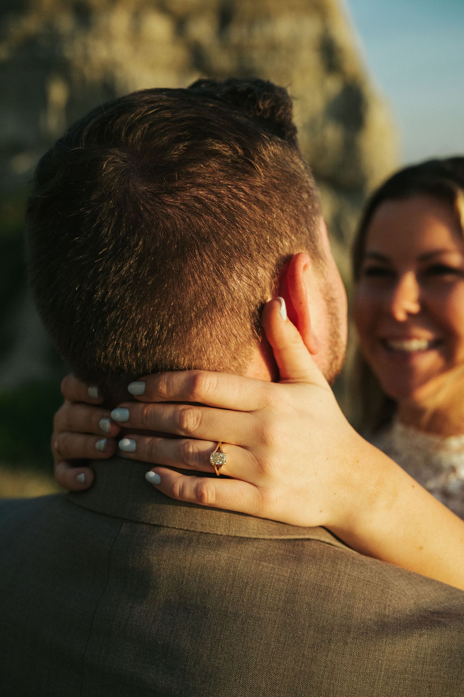 Smiling bride with engagement ring on hand resting on groom's neck during romantic outdoor proposal moment.