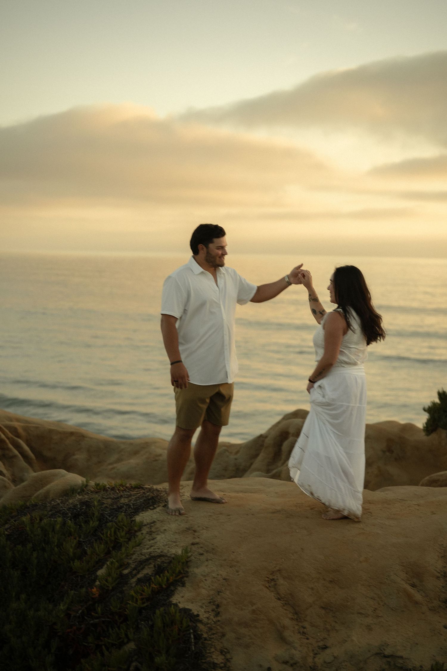 Couple dancing together on rocky coastal cliffs during a warm golden sunset by the ocean.