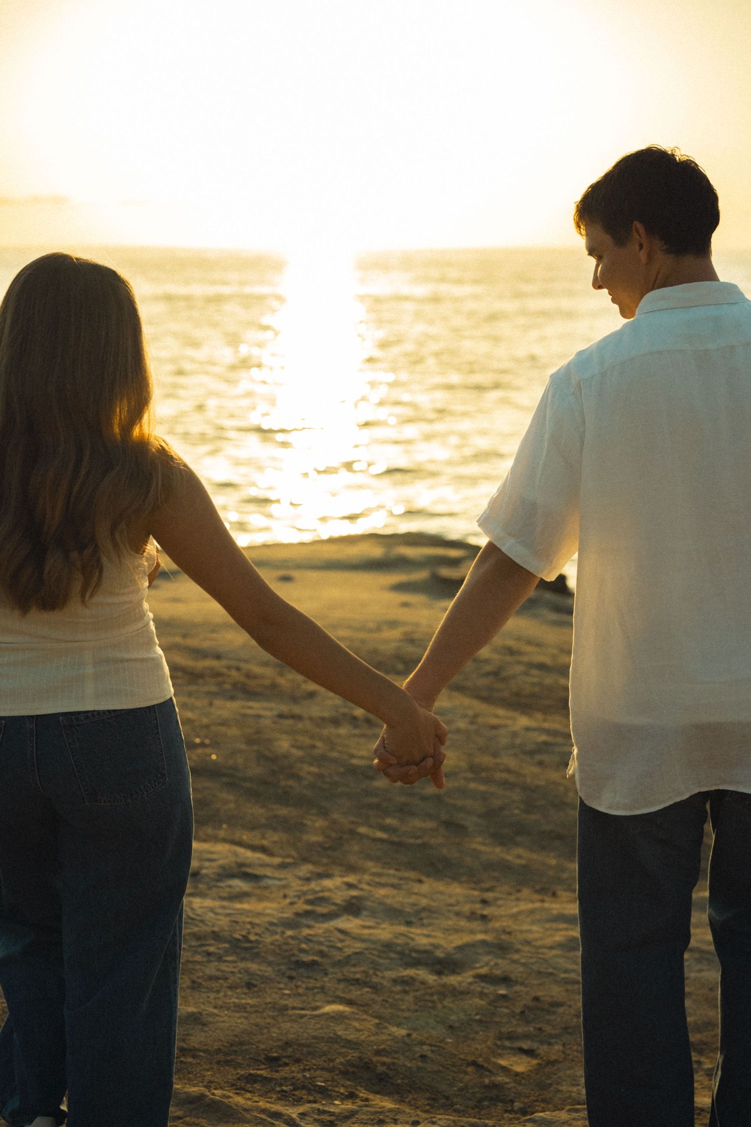 Romantic couple holds hands while watching a golden sunset over the ocean from the beach.