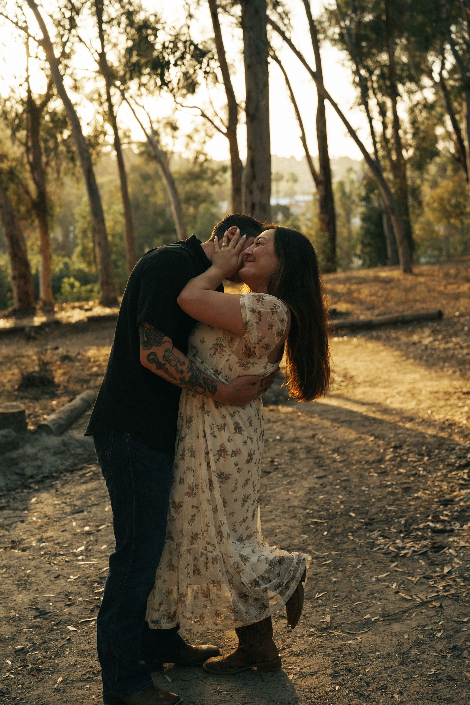 Couple sharing a passionate kiss on a forest path bathed in warm golden sunset light among tall trees.