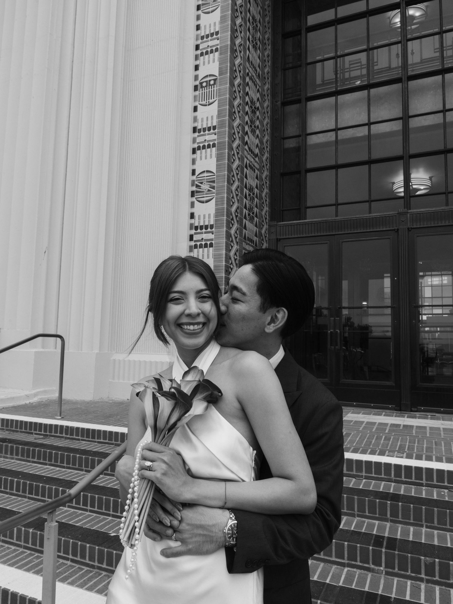 Black and white photo of a couple embracing on steps, woman laughing holding flowers, man kissing her cheek, elegant attire.