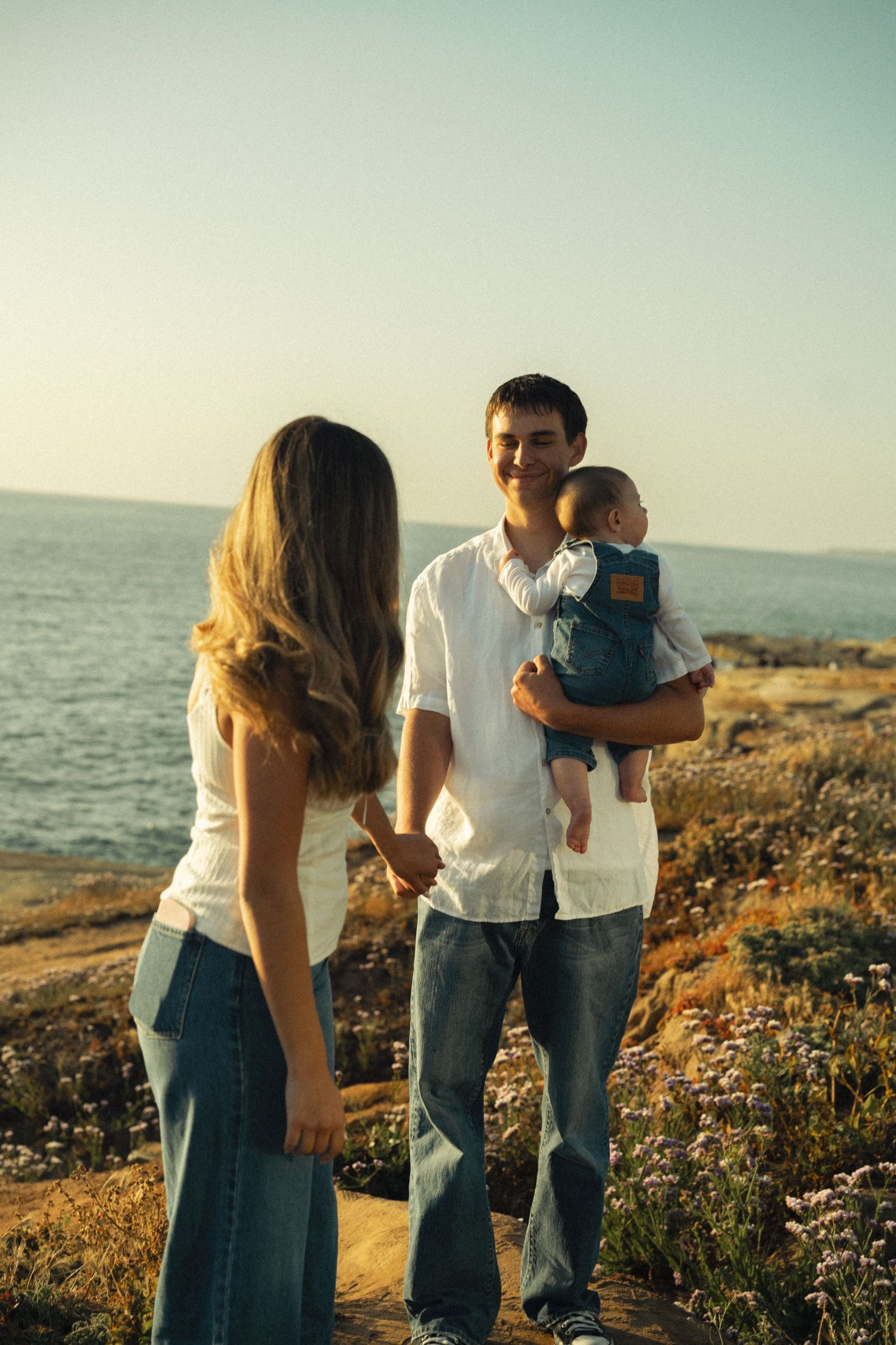 Happy couple holding hands with baby in carrier at scenic coastal overlook during sunset.